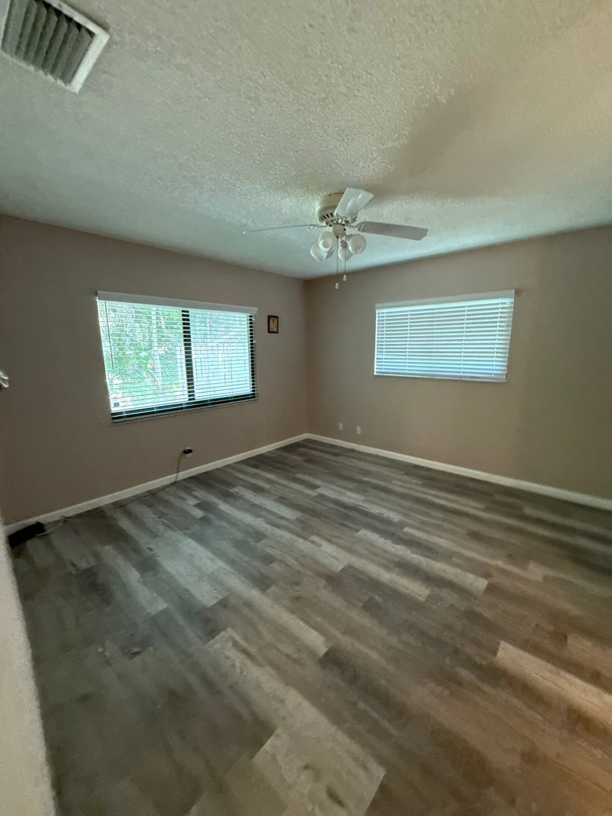 Interior Painting in Boynton Beach, Florida. Empty bedroom with greige painted walls, white baseboards, textured ceiling, ceiling fan, two blinded windows and wood-look plank flooring.