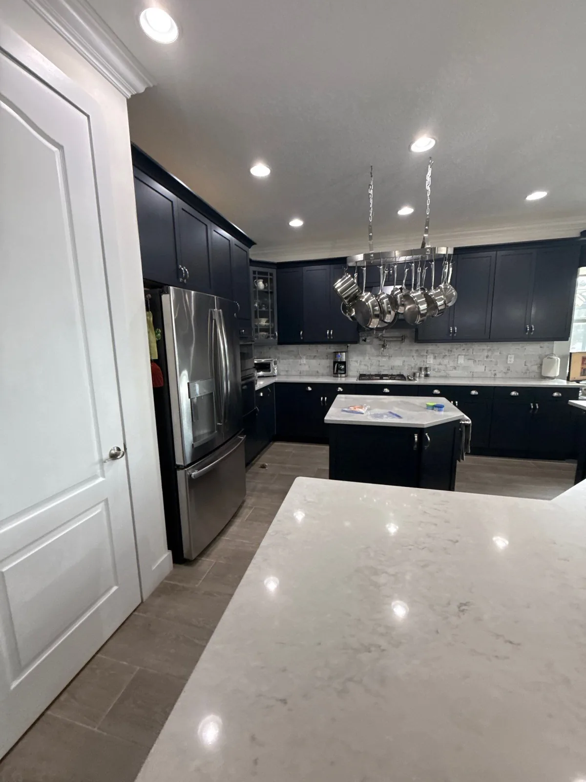 Cabinet Refinishing in Florida Ridge, Florida. Kitchen with dark navy cabinets, stainless steel refrigerator, tile backsplash and white stone island.