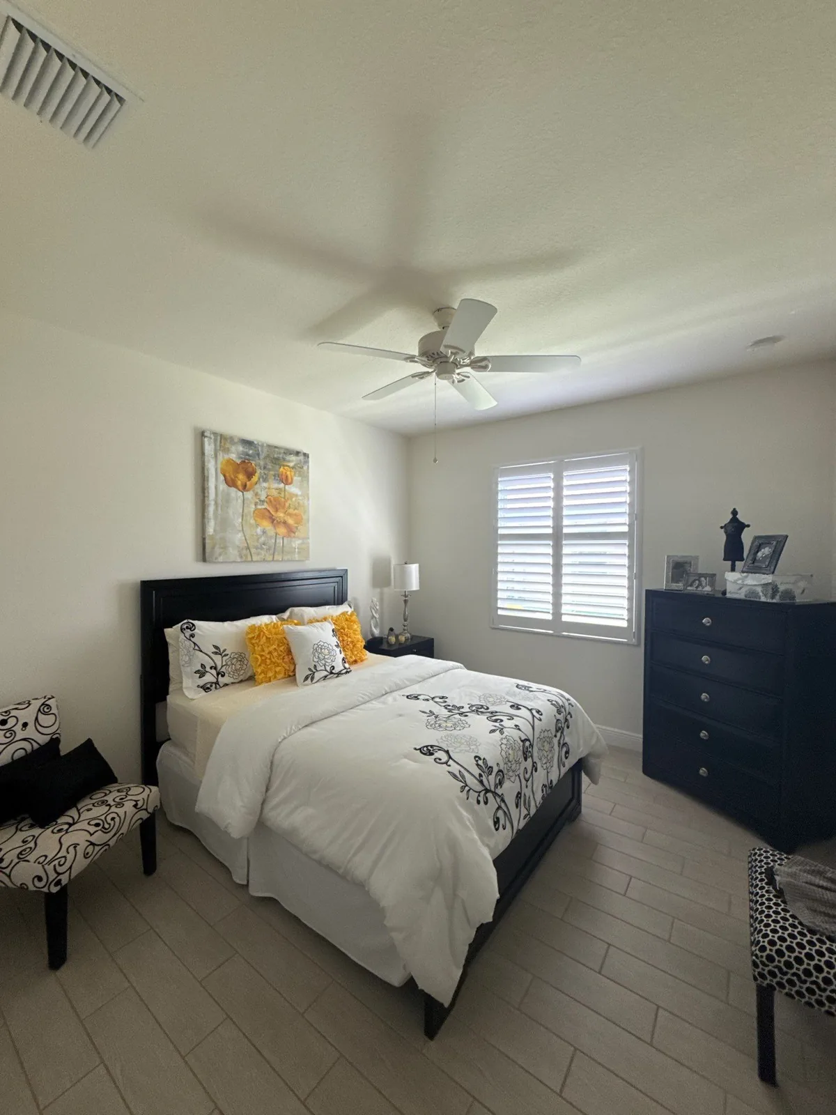 Interior Painting in Florida Ridge, Florida. Staged bedroom with freshly painted walls and ceiling, made bed with dark headboard, ceiling fan, shuttered window and tile floor.