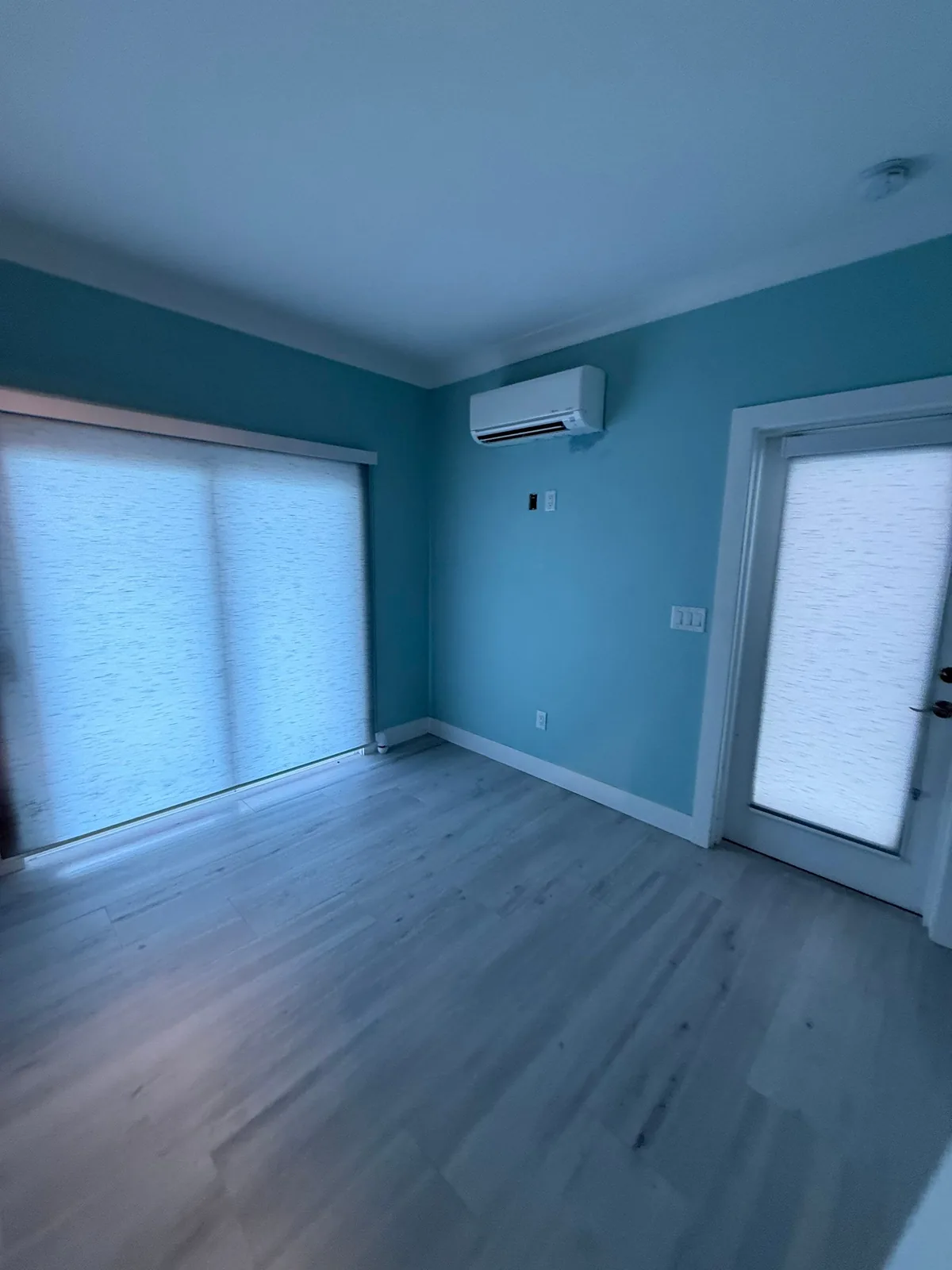 Interior Painting in Fort Pierce, Florida. Empty bedroom with teal walls, white baseboard and door trim, wall-mounted mini-split and grey plank flooring.