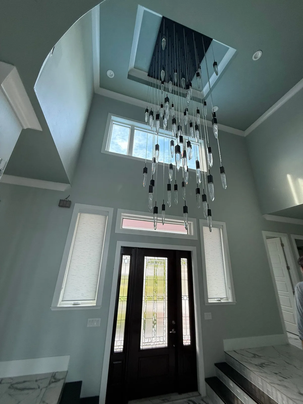 Interior Painting in Fort Pierce, Florida. Two-storey entry foyer with pale blue-grey walls, white crown moulding, a dark front door with sidelights and a hanging linear chandelier.