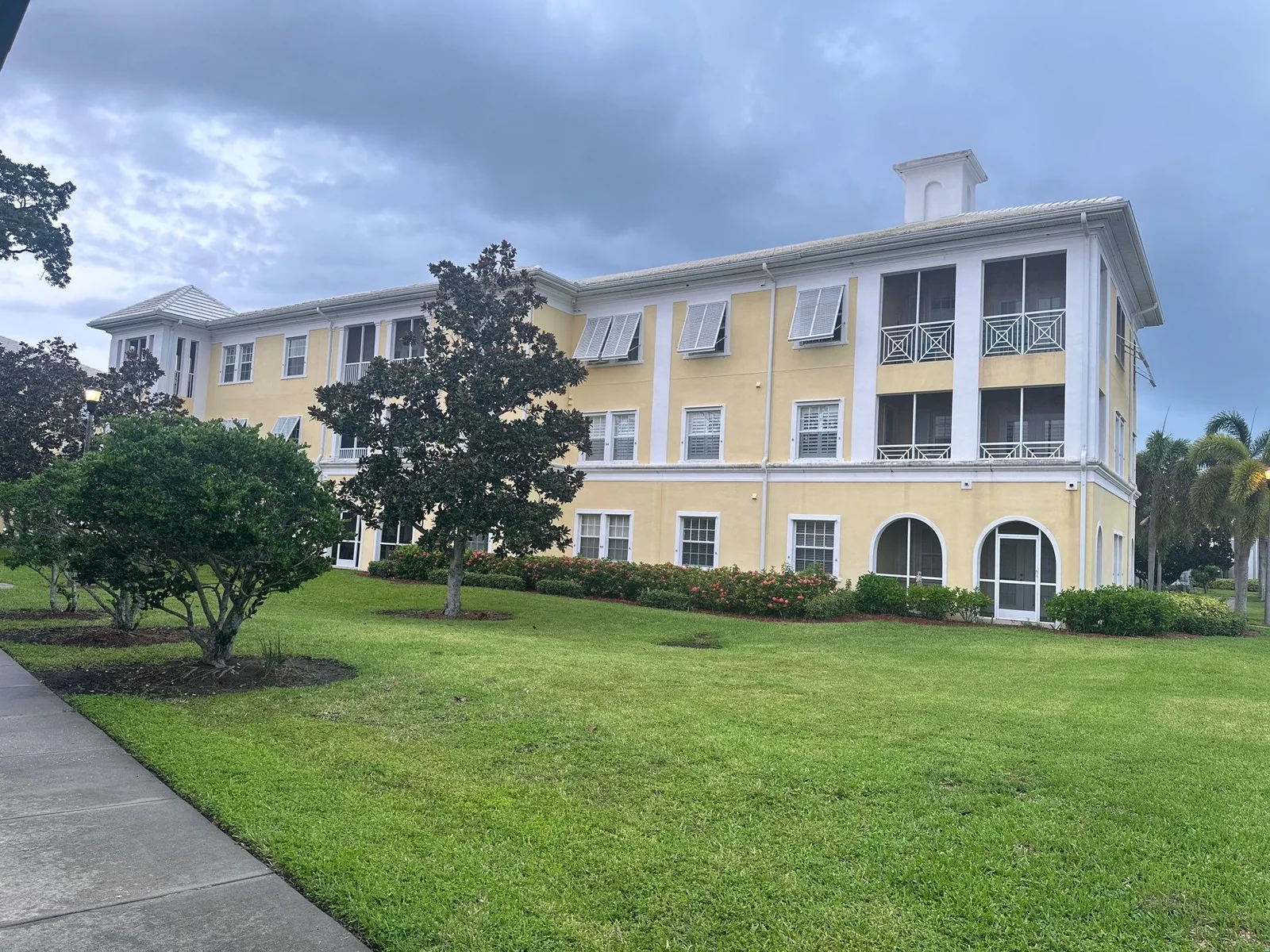 Exterior Painting in Gifford, Florida. Three-storey multi-unit building with yellow exterior walls, white trim, hurricane shutters and screened balconies, seen across a lawn.