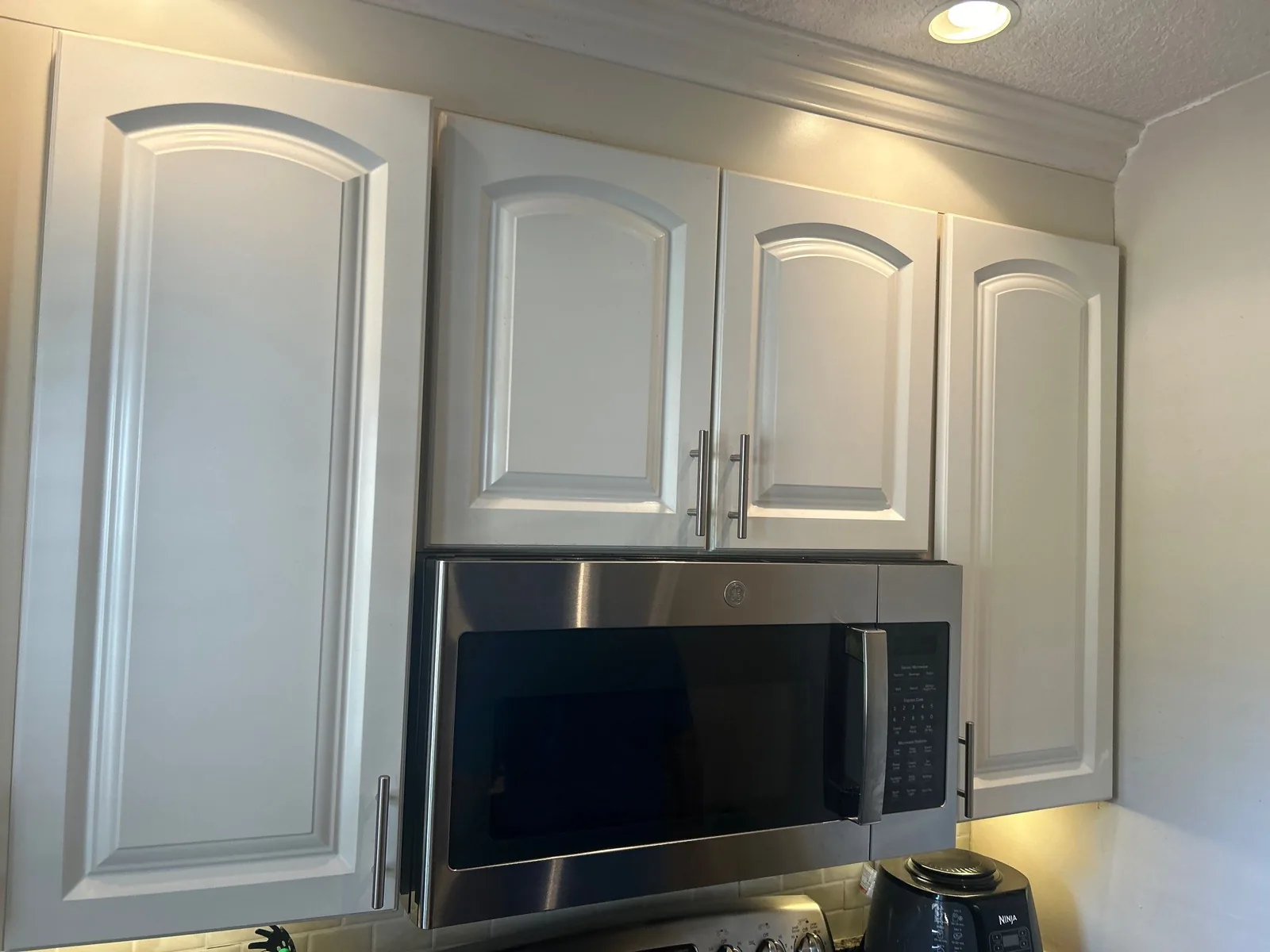 Cabinet Refinishing in Golden Lakes, Florida. White refinished upper cabinets with arched raised-panel doors and bar pulls flanking a stainless over-range microwave.