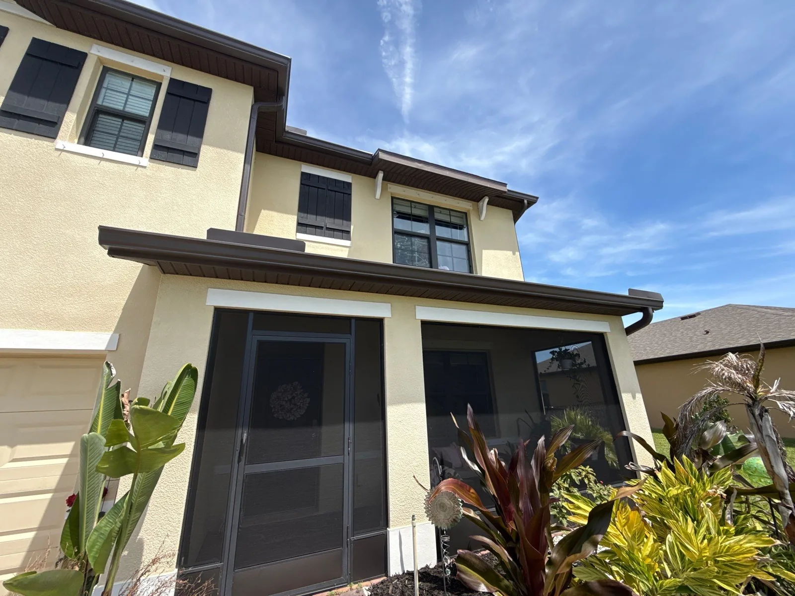 Exterior Painting in Grant-Valkaria, Florida. Corner of a two-storey stucco townhouse, tan walls with brown trim and shutters, screened lanai, tropical planting and blue sky.