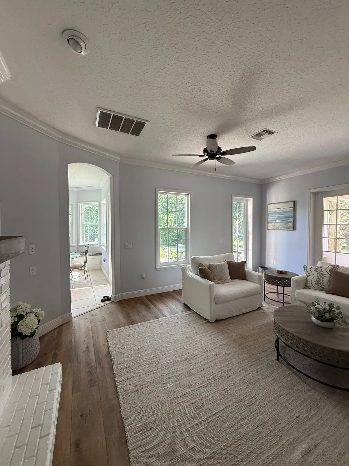 Interior Painting in Grant-Valkaria, Florida. Furnished living room with pale grey walls, white crown moulding and baseboards, a textured ceiling with a ceiling fan, wood-look plank flooring, two white armchairs and a jute area rug.