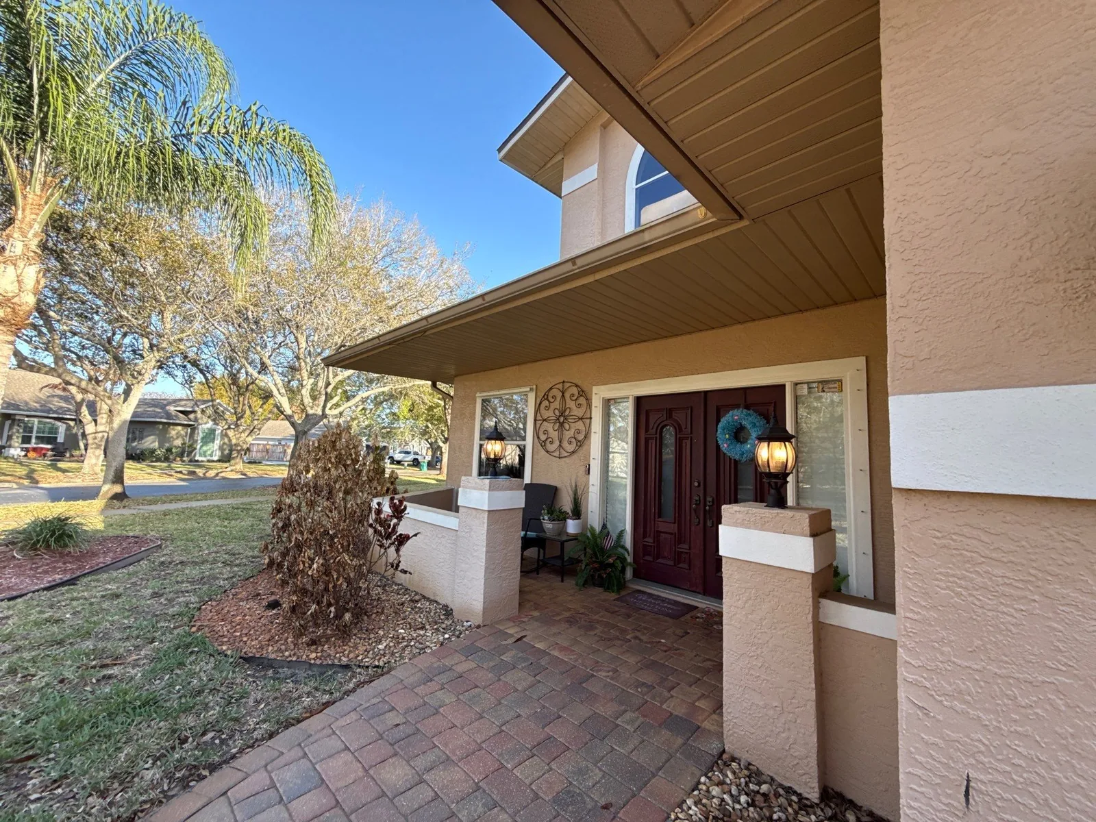 Exterior Painting in Indian Harbour Beach, Florida. Front entry of a two-storey tan stucco home with white trim, a dark wood front door and a paver walkway.