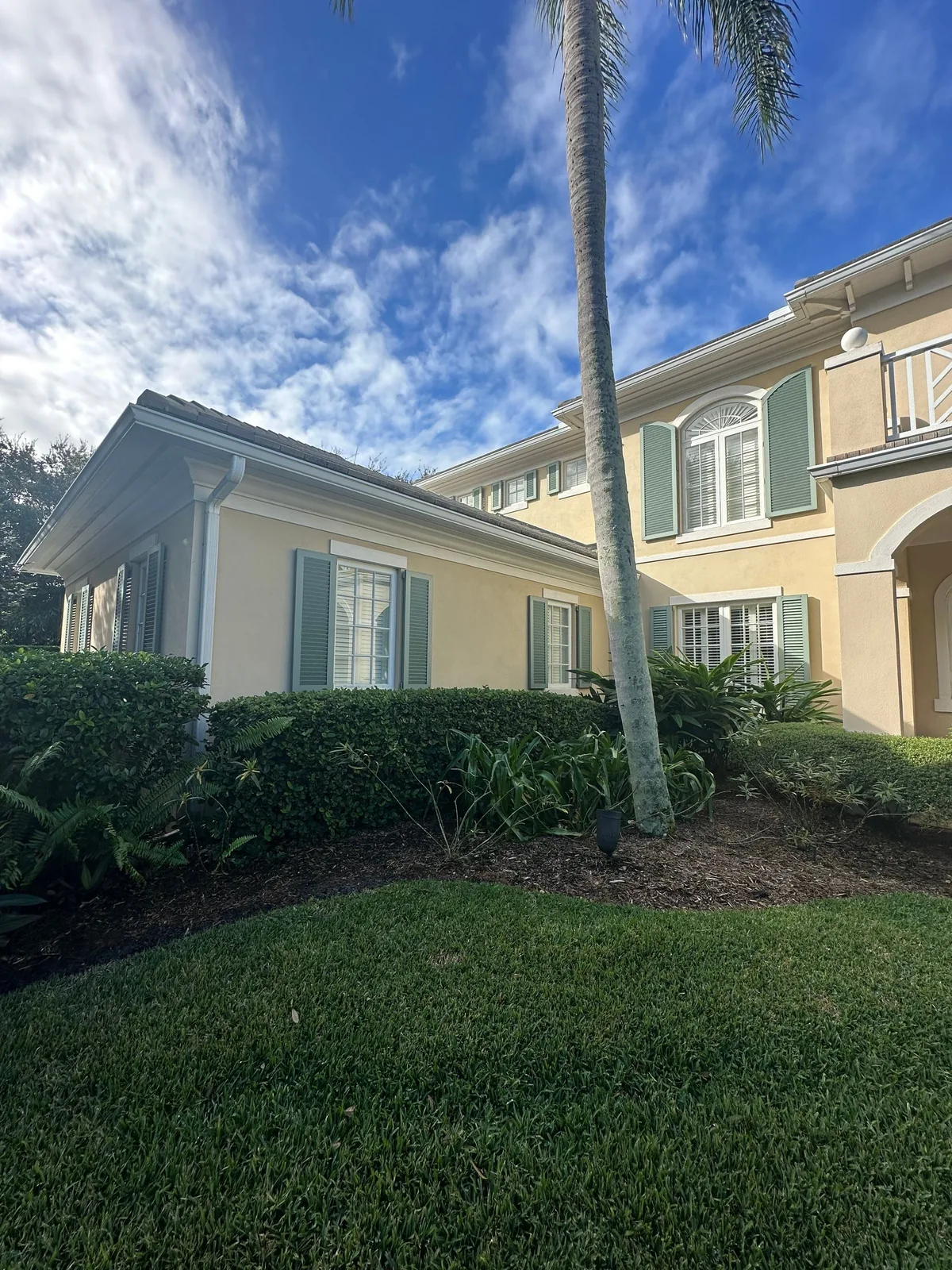 Exterior Painting in Indian River Shores, Florida. Two-storey stucco home in pale yellow with white trim and green louvered shutters, palm tree and hedges in front.
