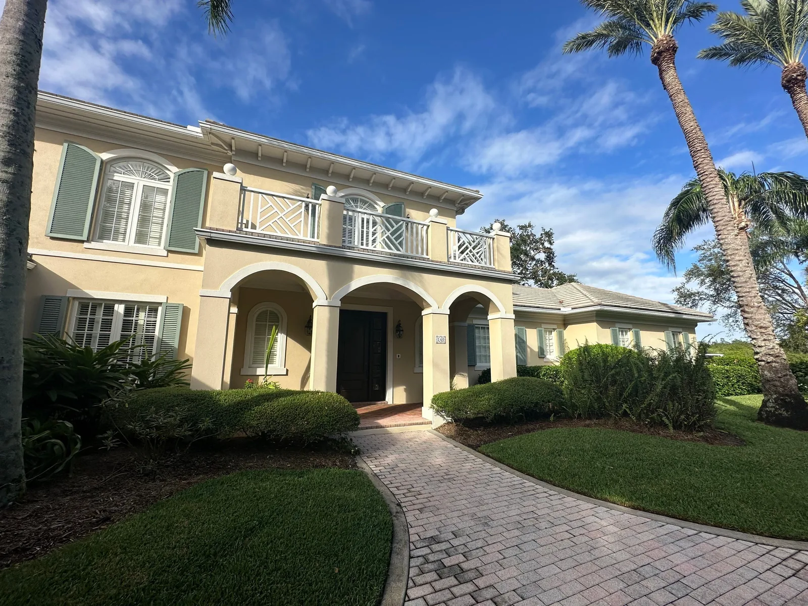 Exterior Painting in Indian River Shores, Florida. Two-storey stucco home in pale yellow with white trim, green shutters, an arched entry and a second-floor balcony, with a paver walkway and lawn in front.