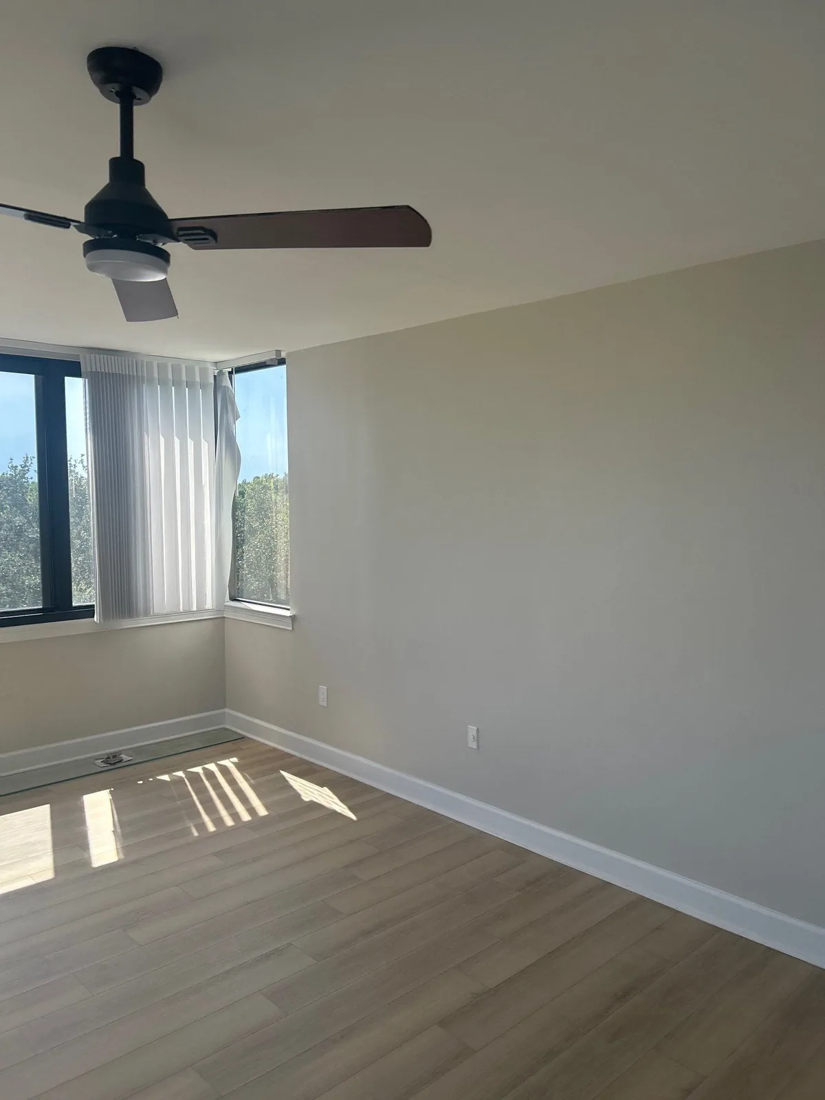 Interior Painting in Indian River Shores, Florida. Empty bedroom with light grey walls, white ceiling, dark ceiling fan, corner windows with vertical blinds and wood-look plank flooring.