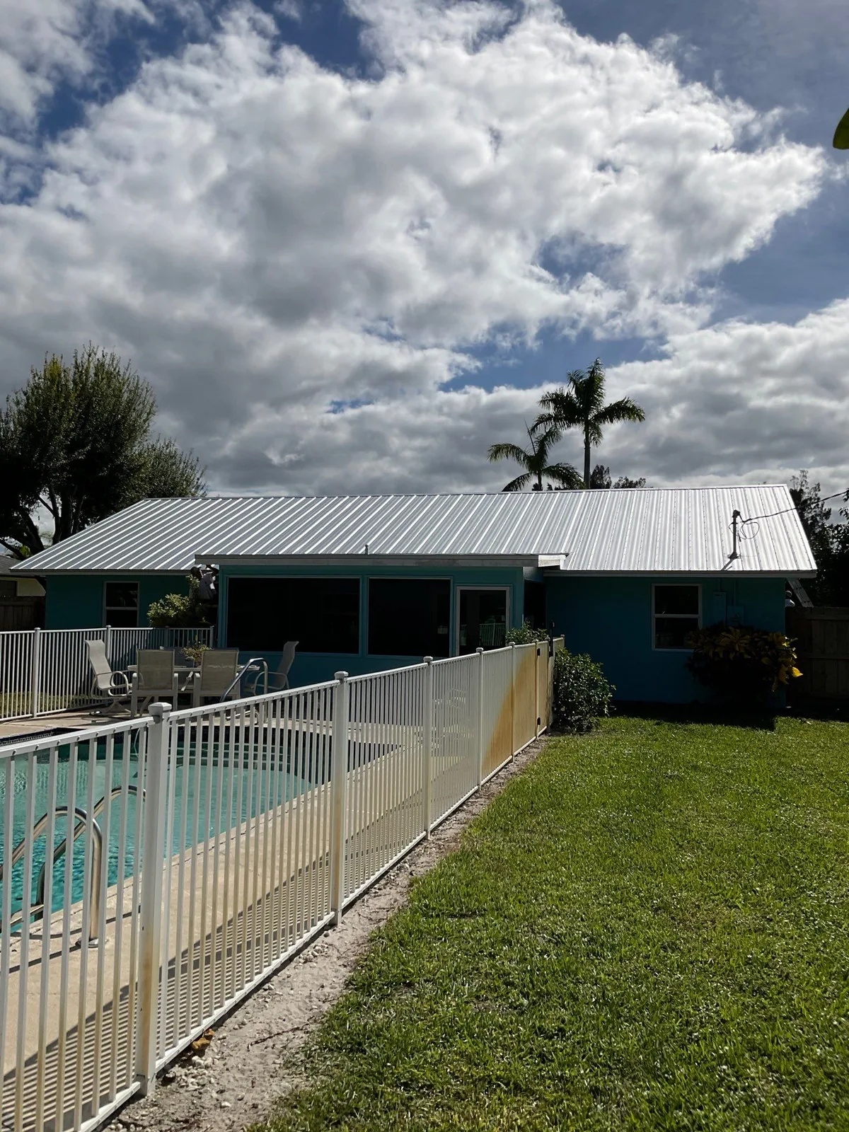 Exterior Painting in Jensen Beach, Florida. Single-storey teal house with white trim and metal roof, seen across a fenced pool and lawn.