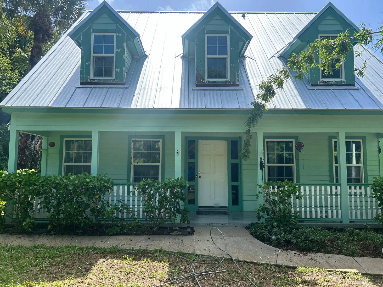 Exterior Painting in Jensen Beach, Florida. Front elevation of a mint green cottage with three dormer windows, white metal roof, covered porch with white railing and a pale entry door.