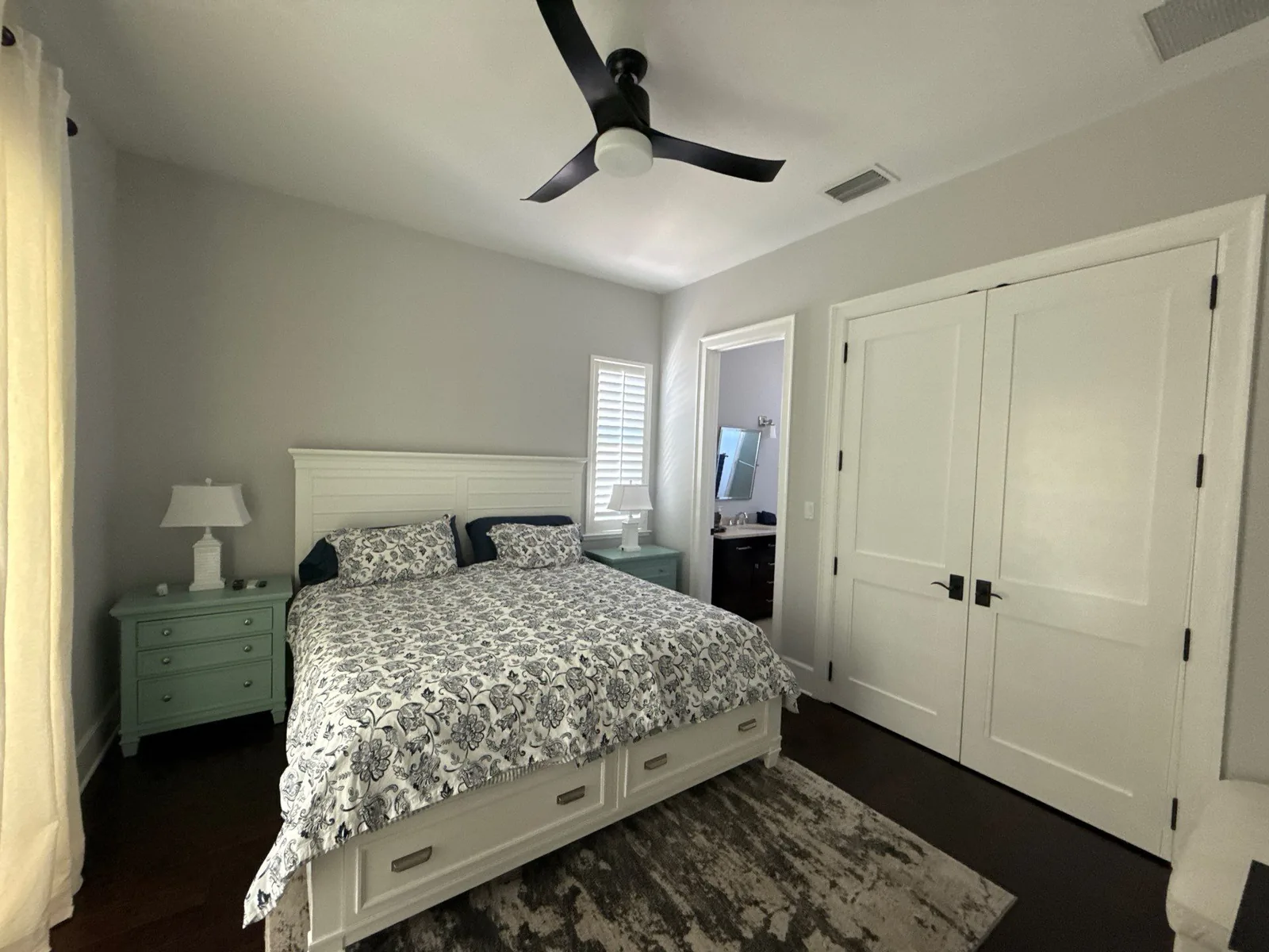 Interior Painting in Jensen Beach, Florida. Bedroom with light grey walls, white-painted double closet doors, white trim and baseboards, made bed and ceiling fan.