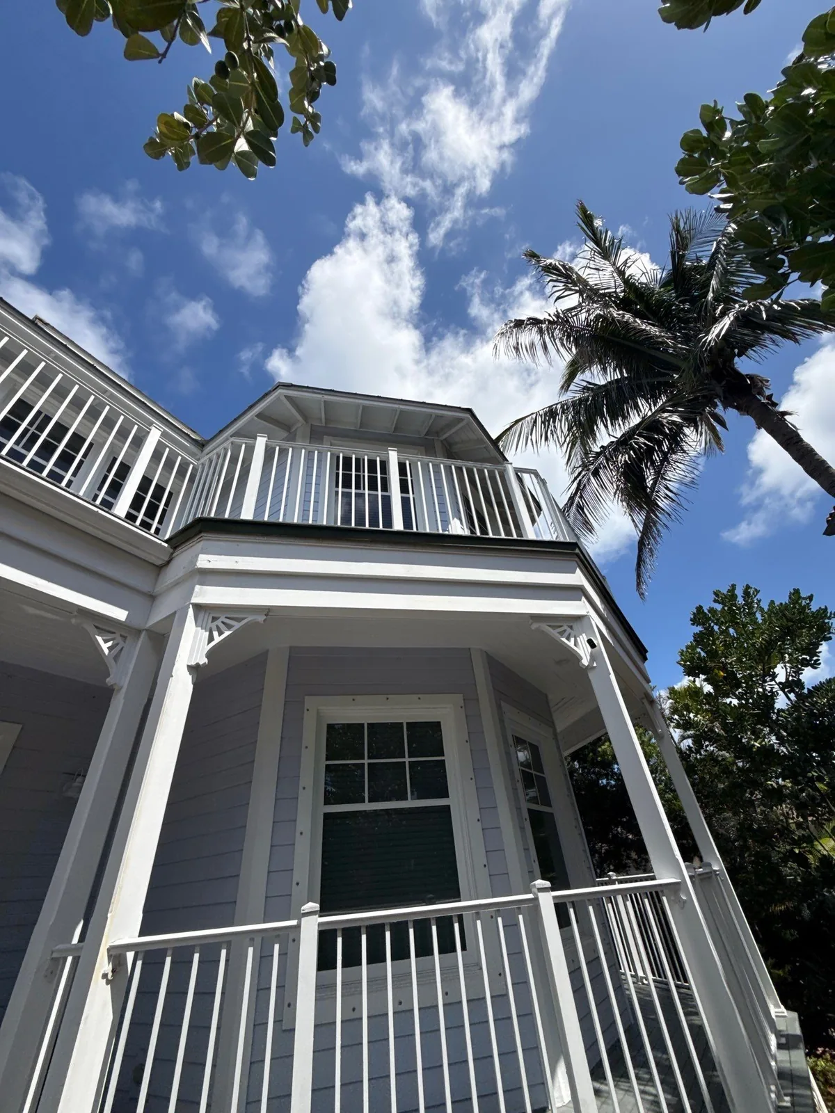 Exterior Painting in Juno Beach, Florida. Two-storey home with pale grey lap siding, white trim, white porch and balcony railings, palm and blue sky.