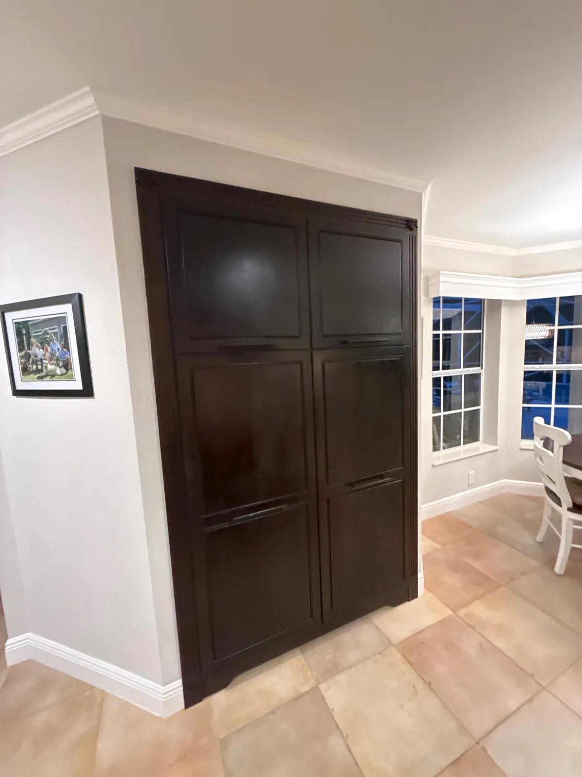 Cabinet Refinishing in Jupiter, Florida. Dark espresso-finished built-in double pantry doors set into a white painted wall with crown moulding and baseboard, beside a tiled floor and bay window.