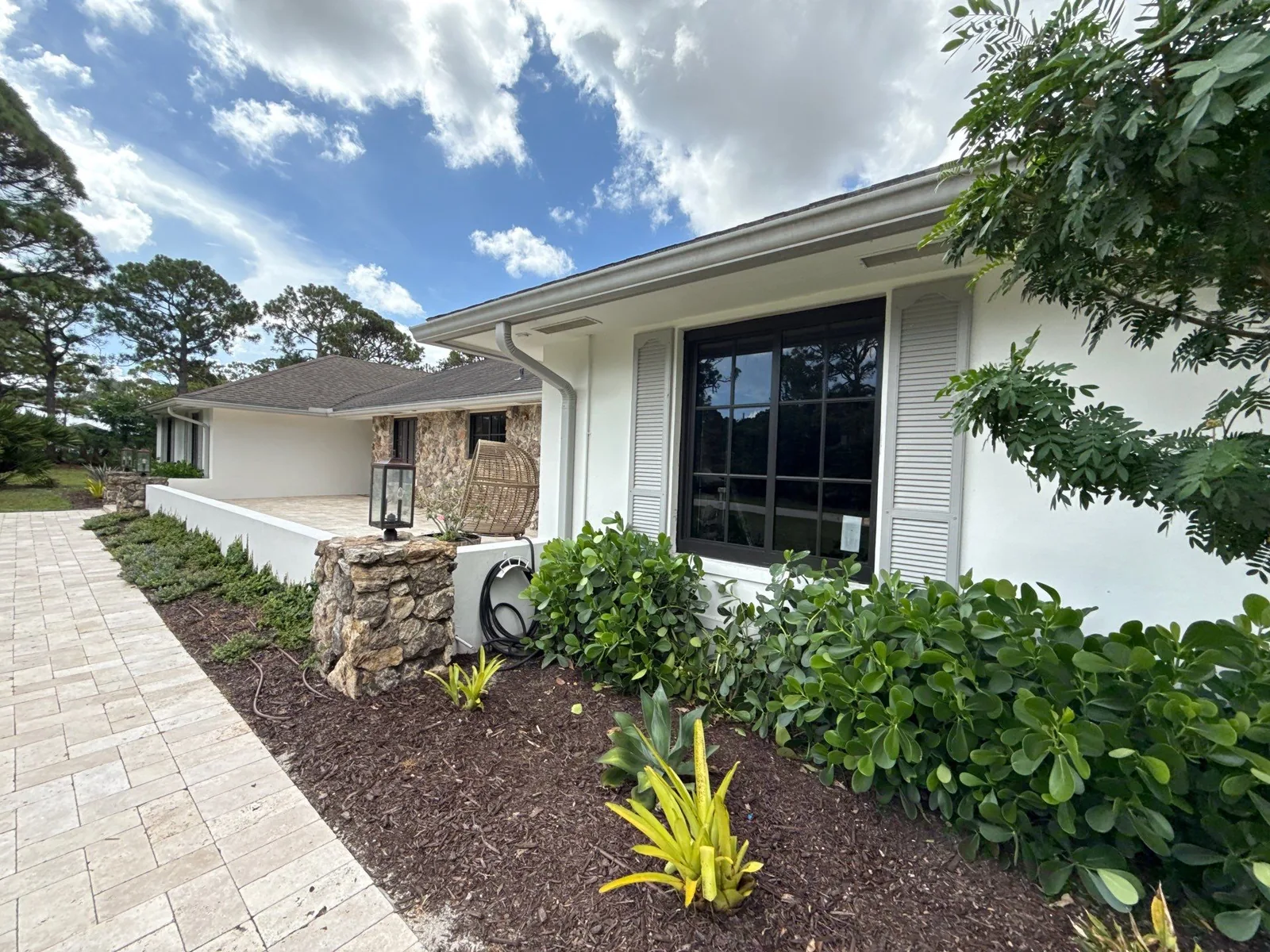 Exterior Painting in Jupiter, Florida. White stucco house exterior with stone accent wall, large dark window and landscaped bed along a paver walkway.