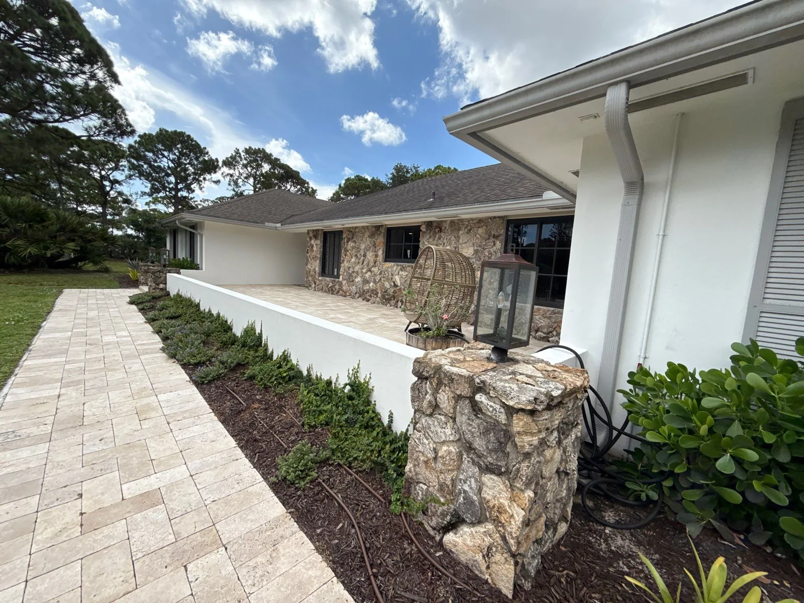 Exterior Painting in Jupiter, Florida. White stucco home exterior with stone pillar and low wall, paver walkway and shrub bed.