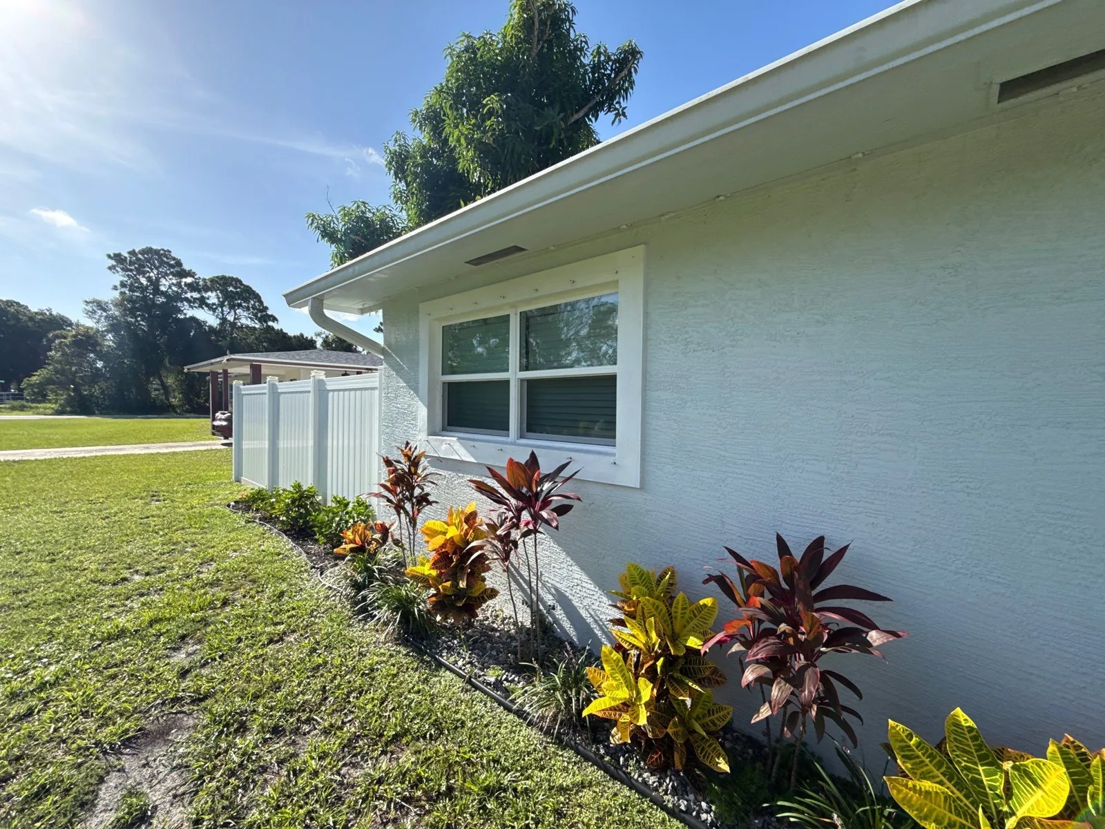 Exterior Painting in Lakewood Park, Florida. Side elevation of a single-storey stucco house painted pale off-white with white fascia, gutter and window trim, and a bed of croton shrubs along the wall.