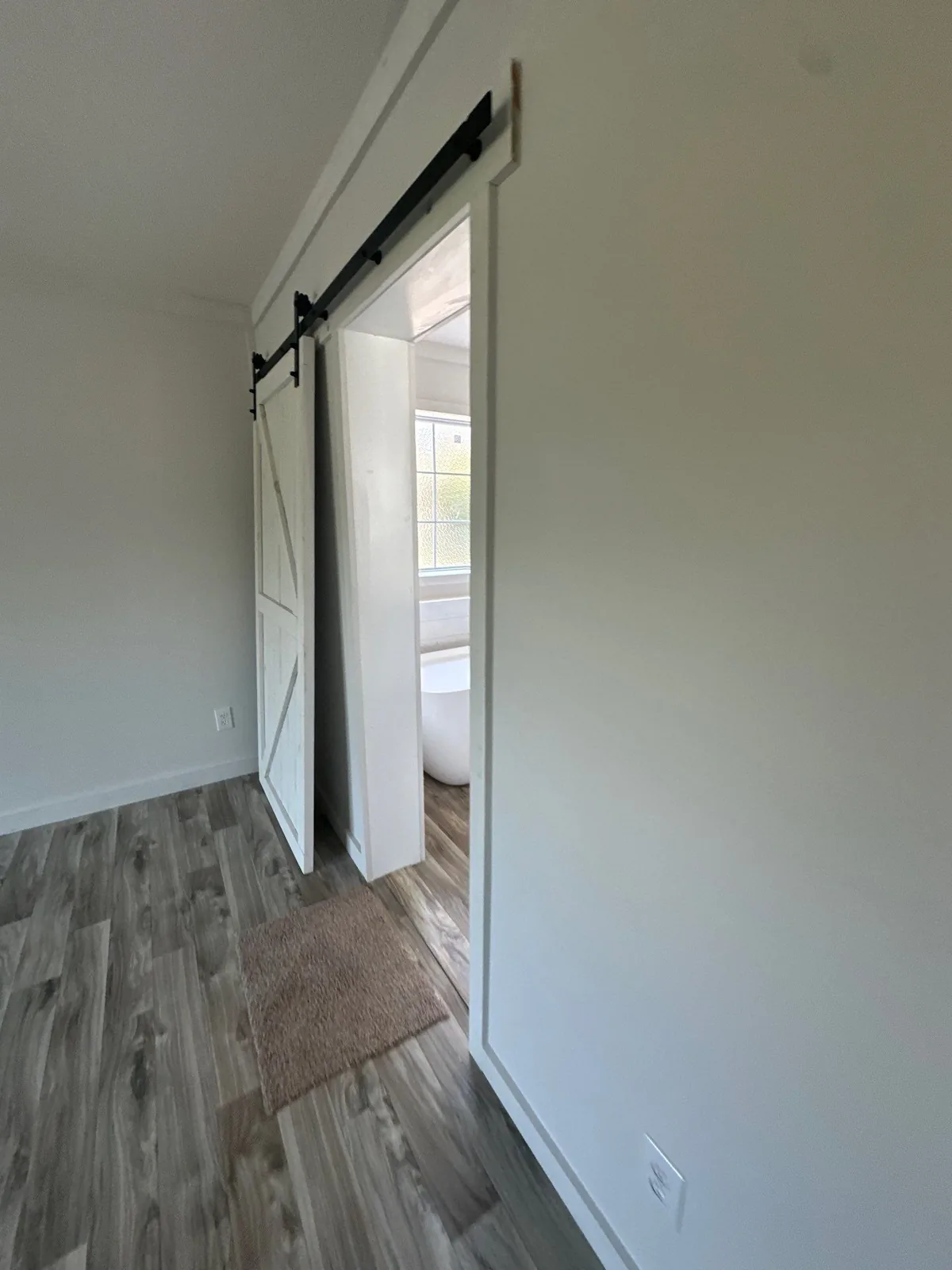 Interior Painting in Melbourne, Florida. White barn door on a black rail opening to a bathroom, with white walls and wood-look plank flooring.
