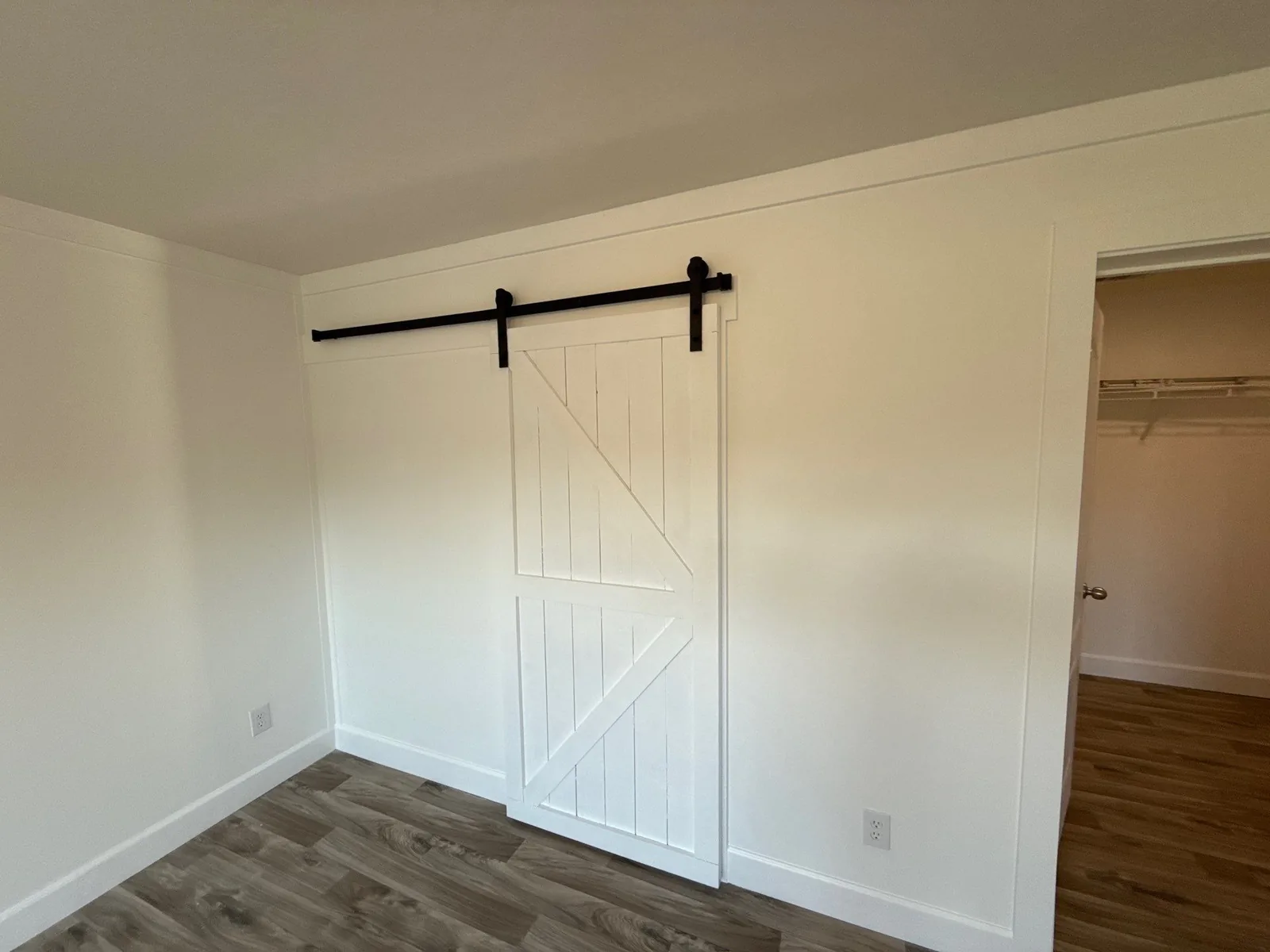 Interior Painting in Melbourne, Florida. Bedroom with white painted walls and baseboards, a white barn door on a black sliding track, and grey wood-look plank flooring.