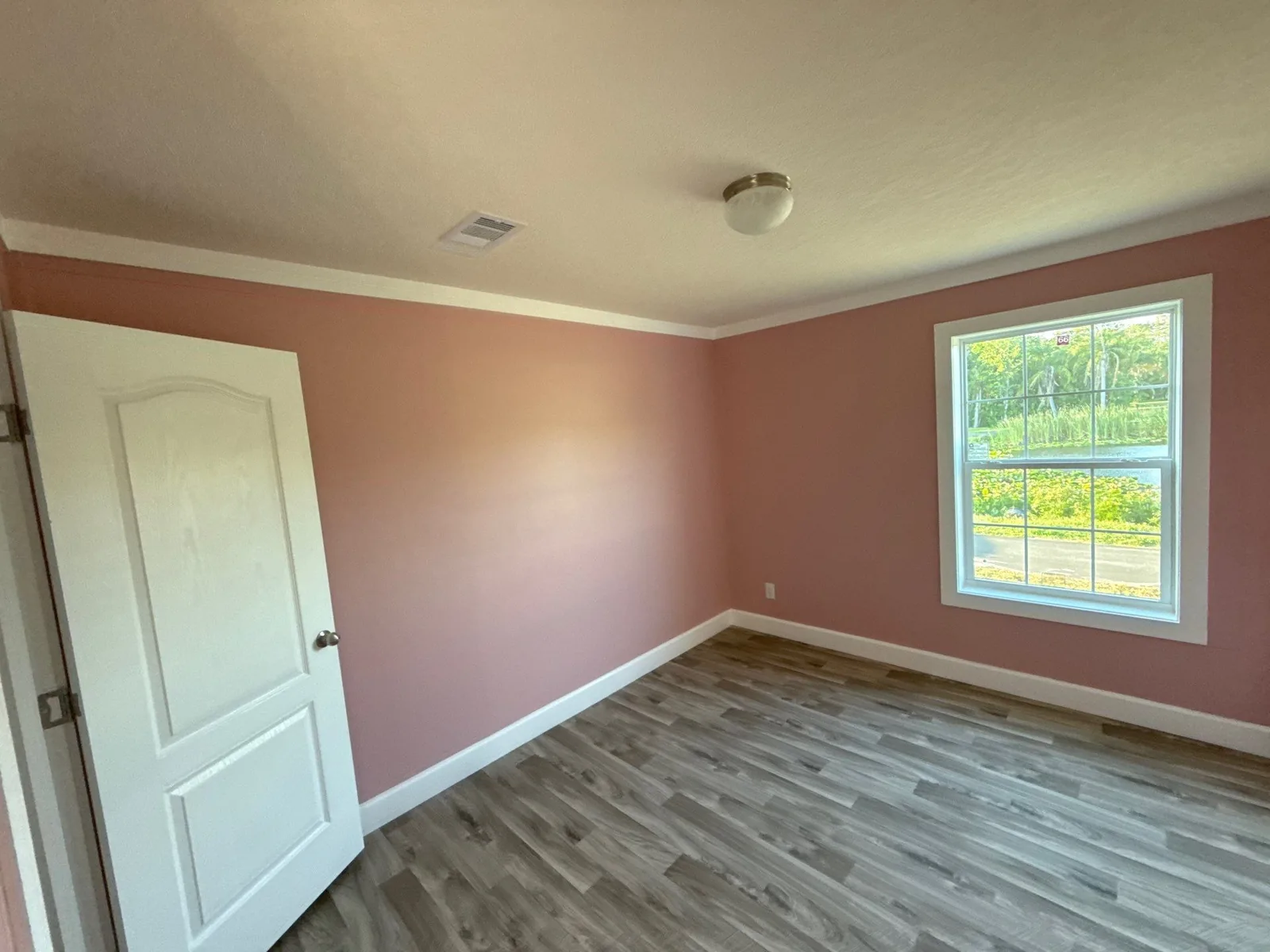 Interior Painting in Melbourne, Florida. Empty bedroom with dusty rose painted walls, white door and trim, and wood-look vinyl flooring.