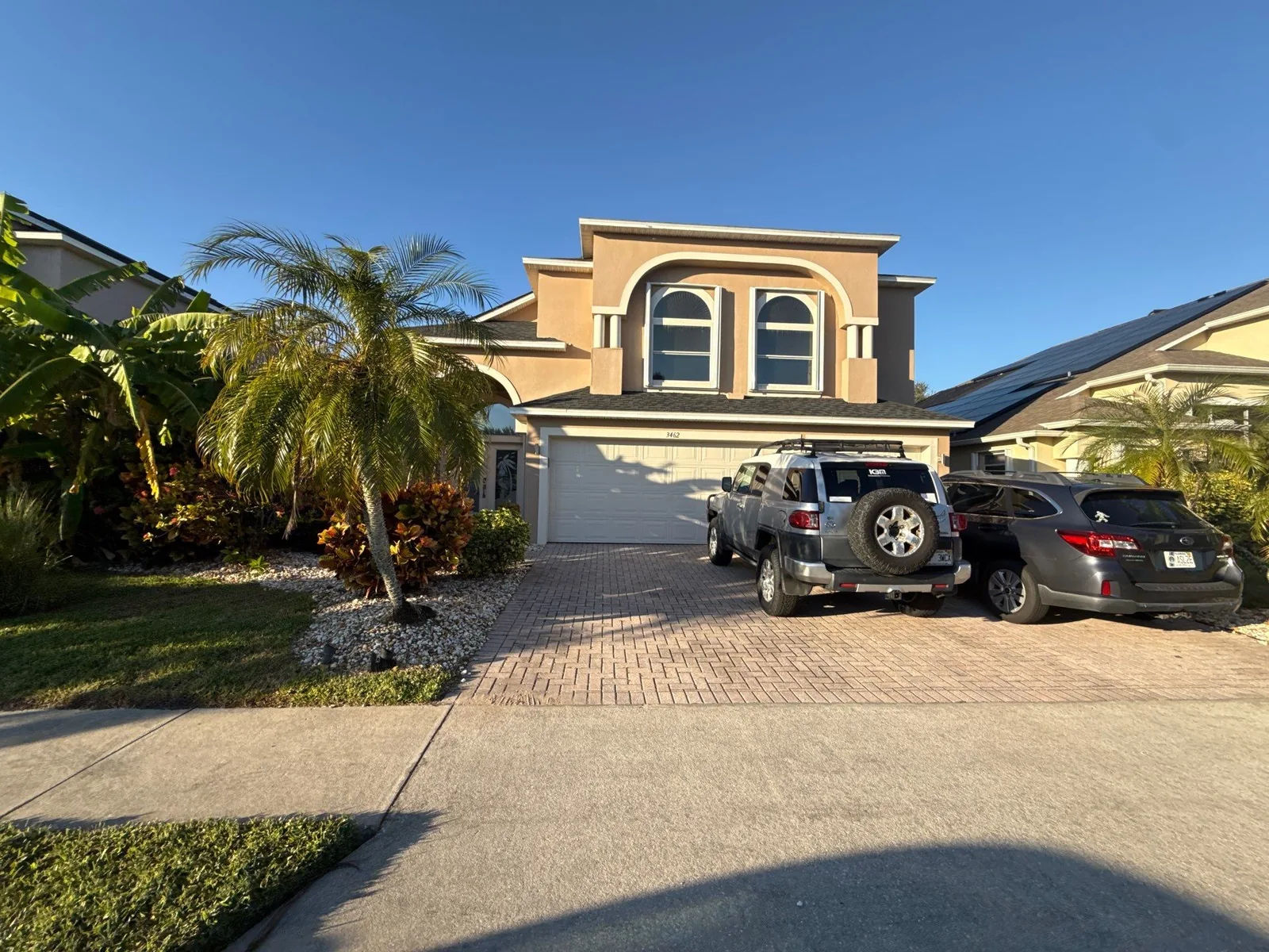 Exterior Painting in Merritt Island, Florida. Two-storey stucco home with arched upper window, white garage door and paver driveway with parked vehicles.