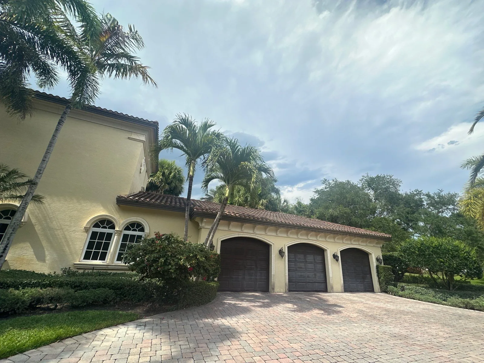 Exterior Painting in North Palm Beach, Florida. Stucco home with three dark garage doors, arched windows, barrel tile roof and paver driveway under a blue sky.