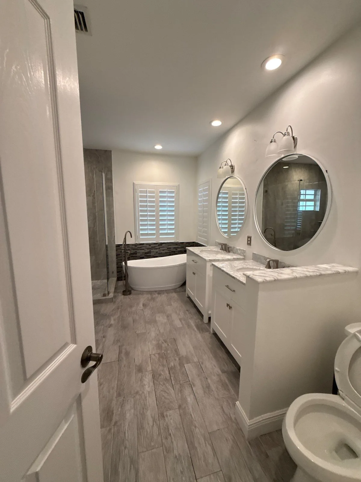 Interior Painting in North Palm Beach, Florida. Bathroom with a freestanding white tub, white vanity with two round mirrors, tiled shower, plantation shutters and wood-look tile flooring.