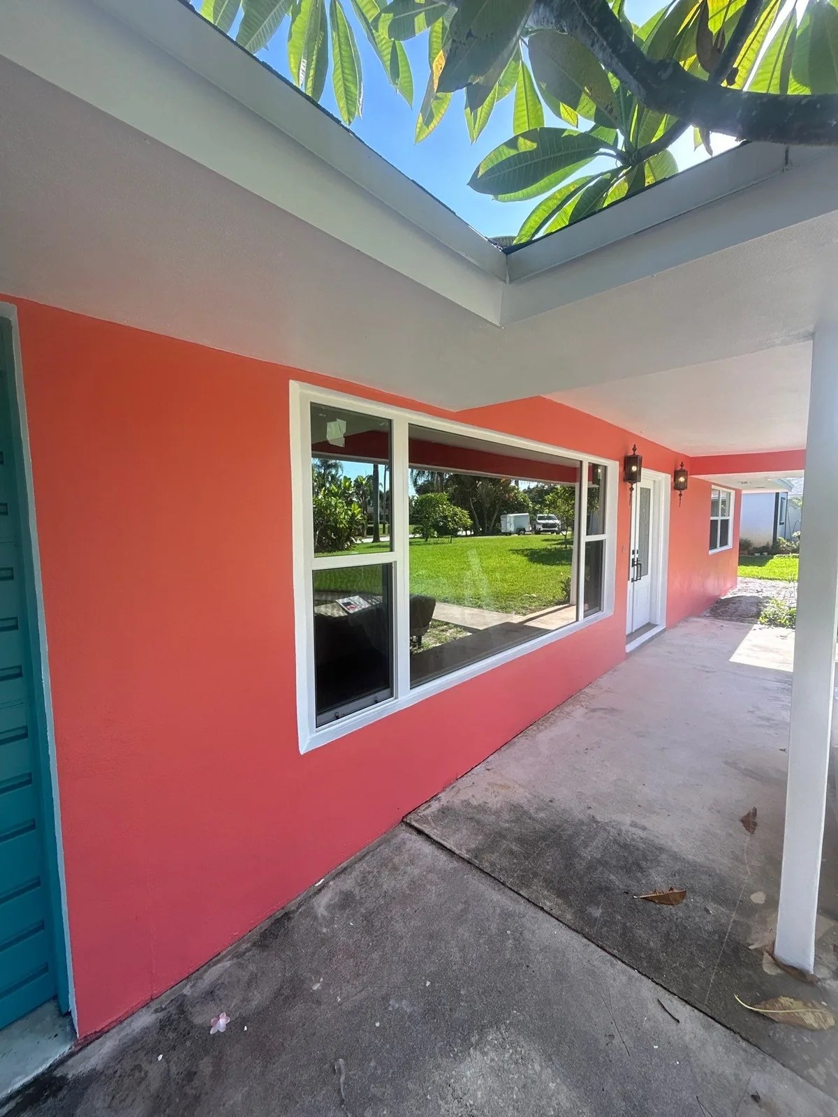 Exterior Painting in North River Shores, Florida. Coral stucco exterior wall with a white-trimmed picture window under a covered walkway, blue sky and palm fronds above.