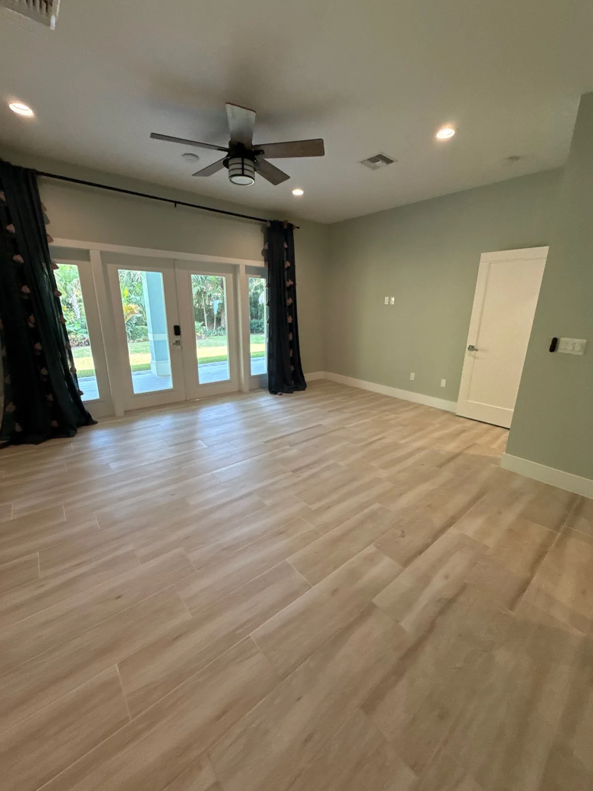 Interior Painting in North River Shores, Florida. Empty living room with sage green walls, white trim and baseboard, French doors with dark curtains and a white interior door.