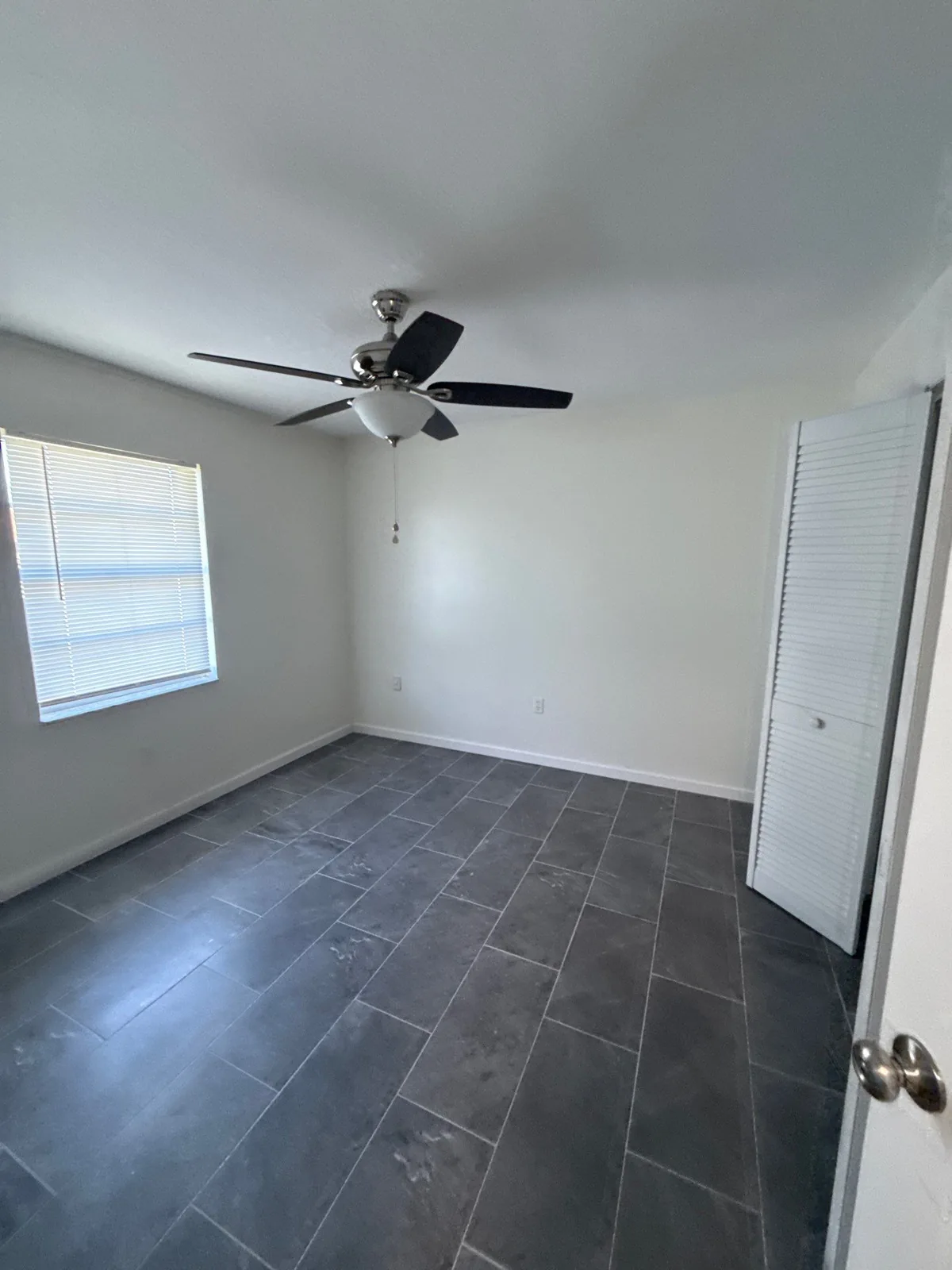 Interior Painting in Palm Bay, Florida. Empty bedroom with freshly painted light walls and white ceiling, a dark ceiling fan, grey tile floor and white louvered closet doors.