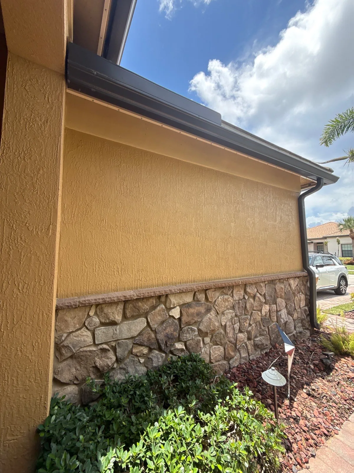 Exterior Painting in Palm City, Florida. Side elevation of a single-storey home with freshly painted gold-toned stucco, brown fascia and soffit, and a stone veneer base.