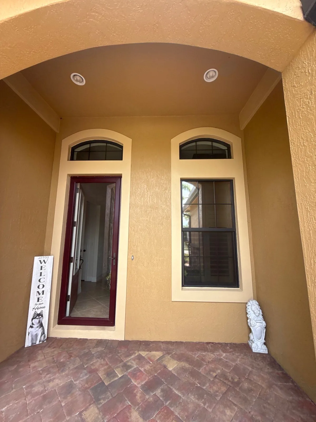 Exterior Painting in Palm City, Florida. Arched front entry with gold-toned stucco walls, a dark red front door and matching dark window trim above a brick paver porch.