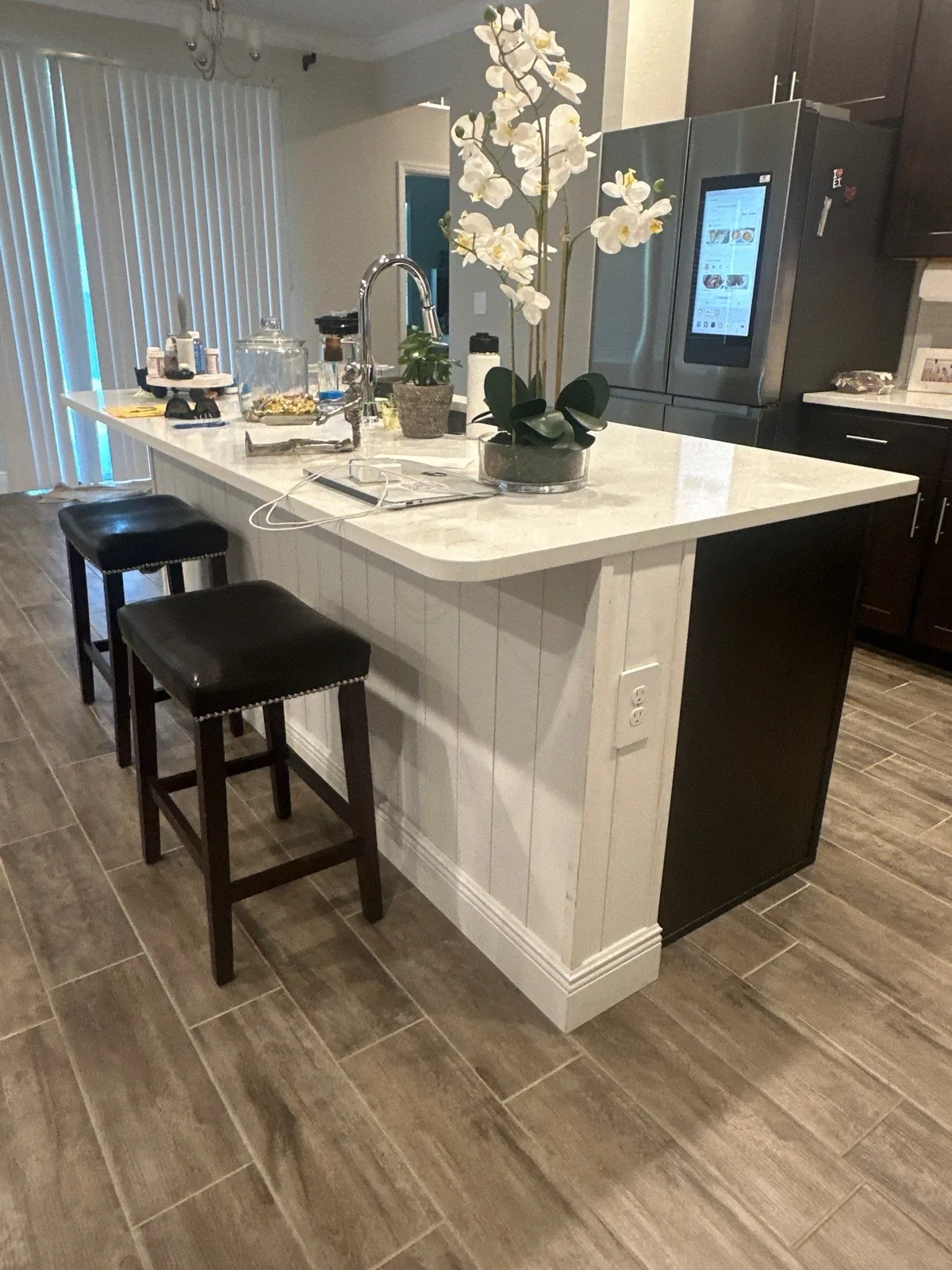 Cabinet Refinishing in Port Saint Lucie, Florida. Kitchen island with a white painted panelled base and light stone countertop, two dark bar stools, with dark cabinetry and a stainless refrigerator behind.