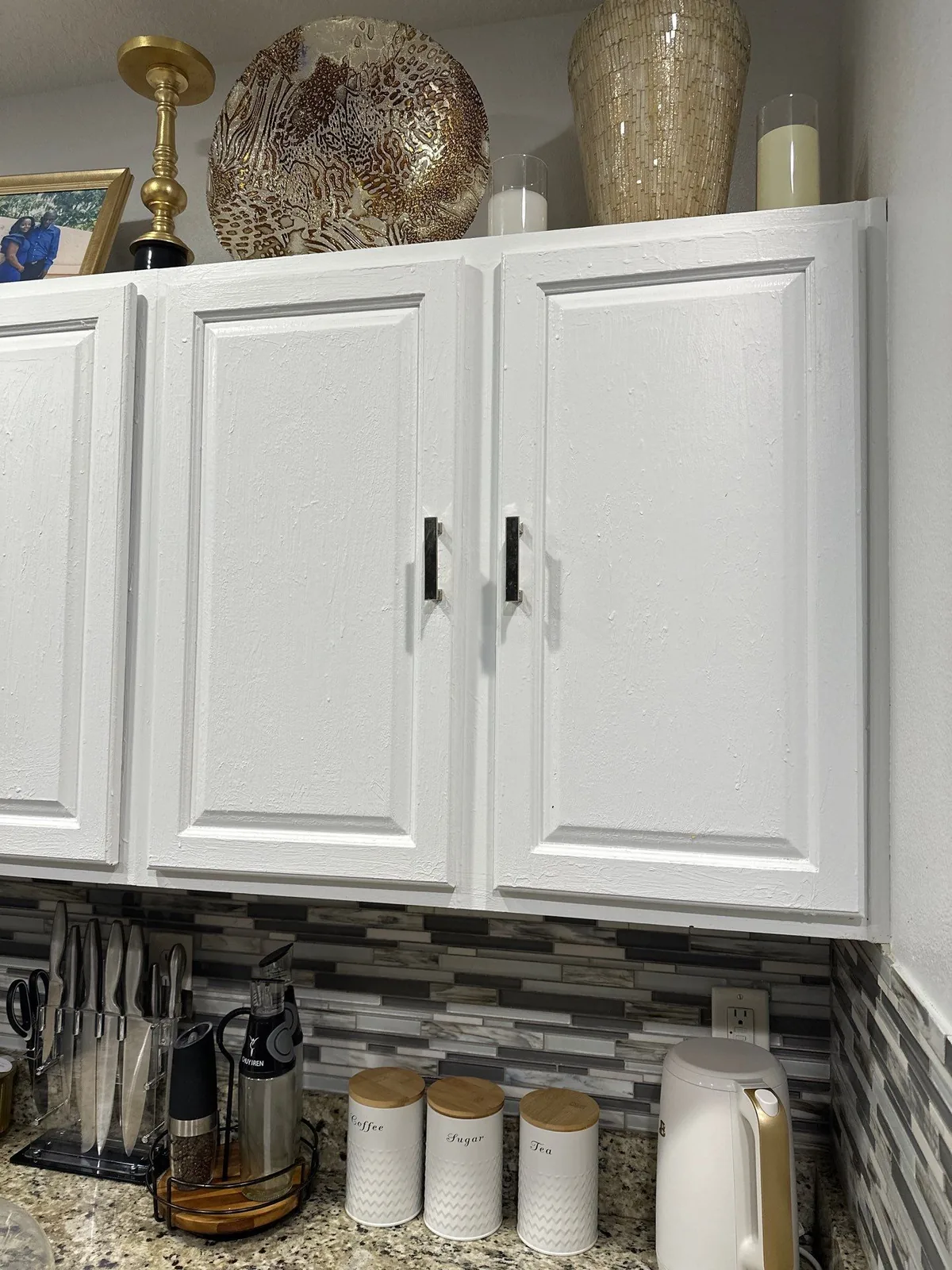 Cabinet Refinishing in Port Saint Lucie, Florida. White-painted upper kitchen cabinets with black handles above a mosaic tile backsplash and granite countertop.