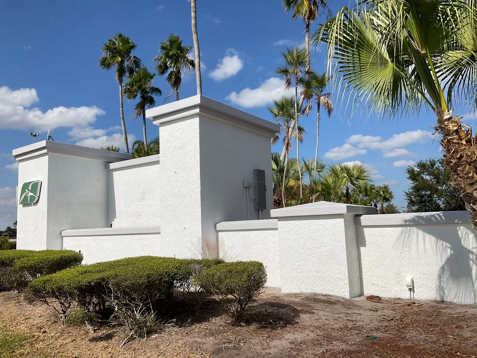 Exterior Painting in Port Saint Lucie, Florida. White painted stucco entrance wall with pillars and a raised emblem, palms and blue sky behind.