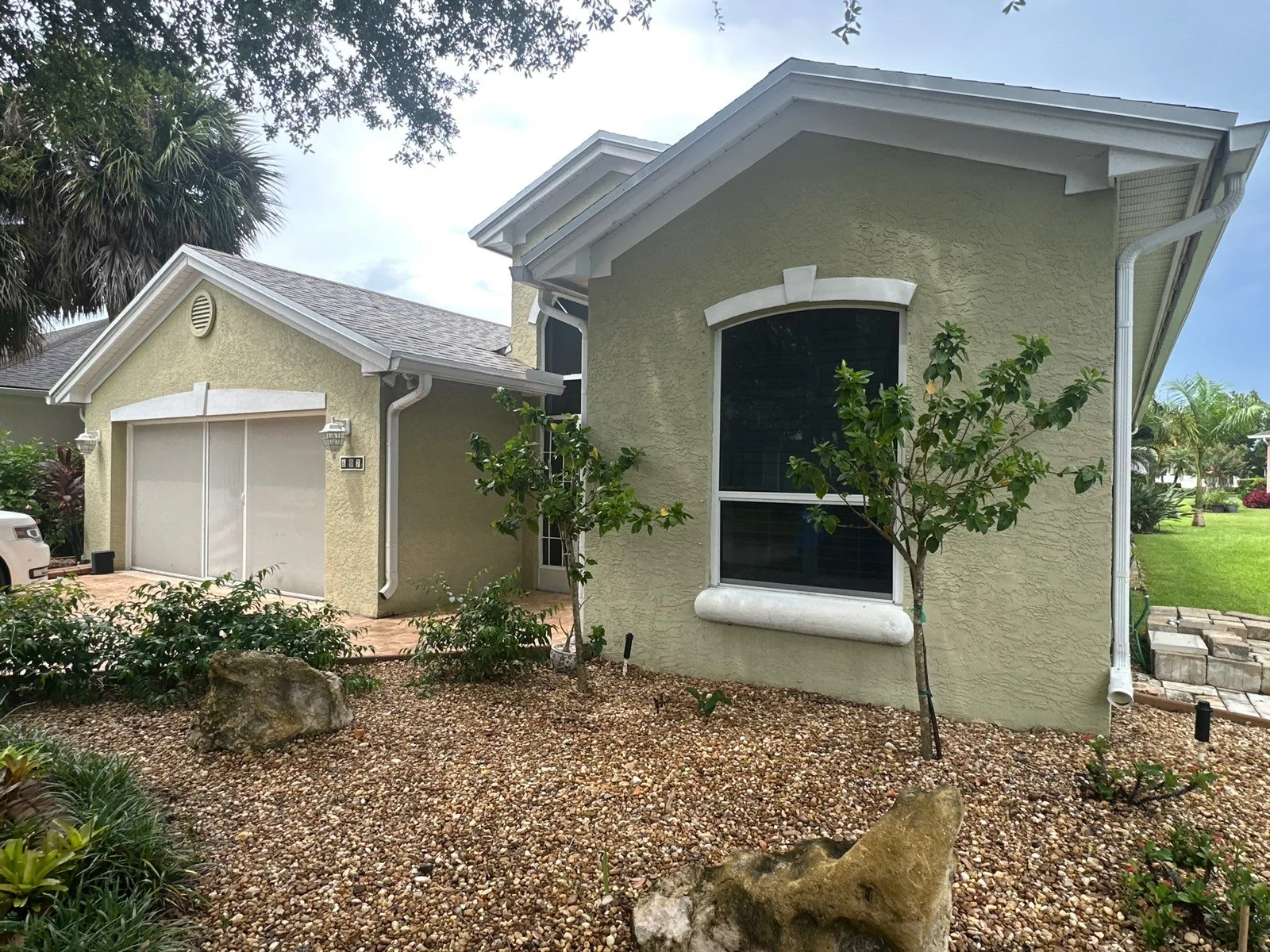 Exterior Painting in Port Saint Lucie, Florida. Front corner of a pale green stucco home with white trim, gabled roofline, arched window surround and attached garage.