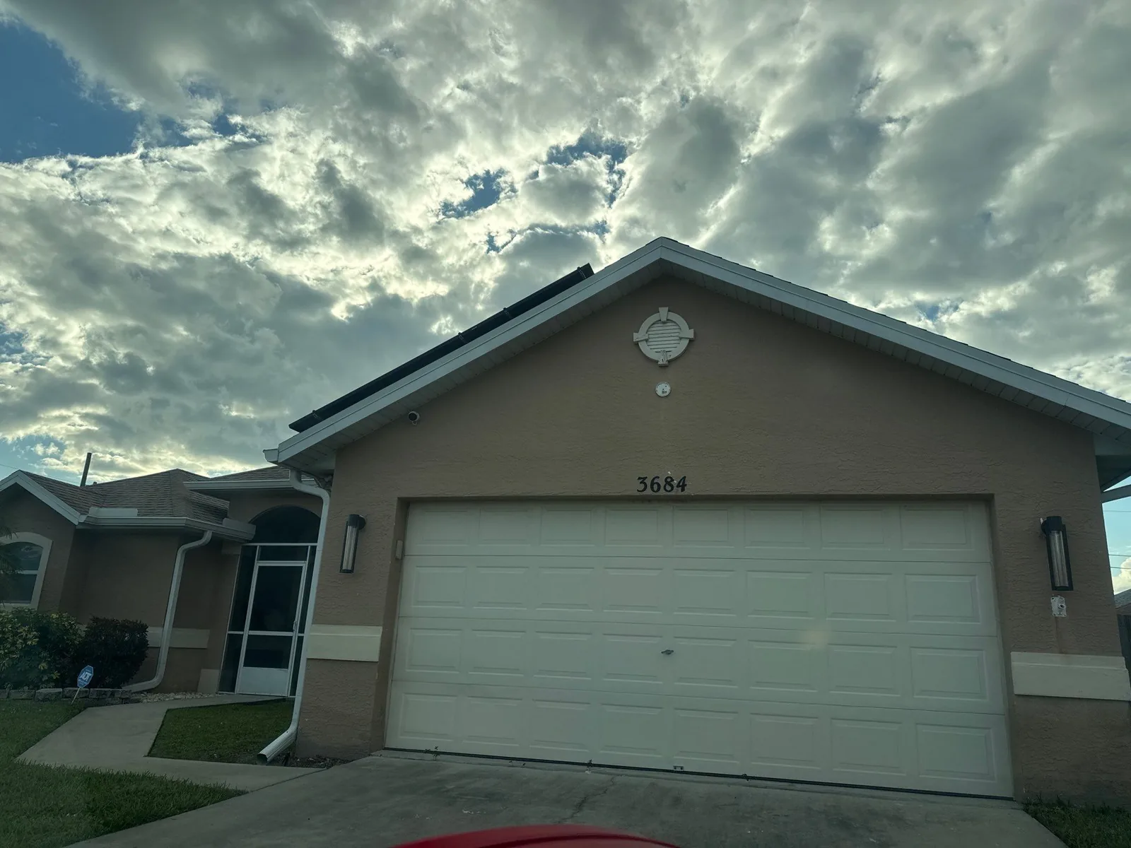 Exterior Painting in Port Saint Lucie, Florida. Front elevation of a single-storey stucco house painted tan with a white two-car garage door, under a cloudy sky.