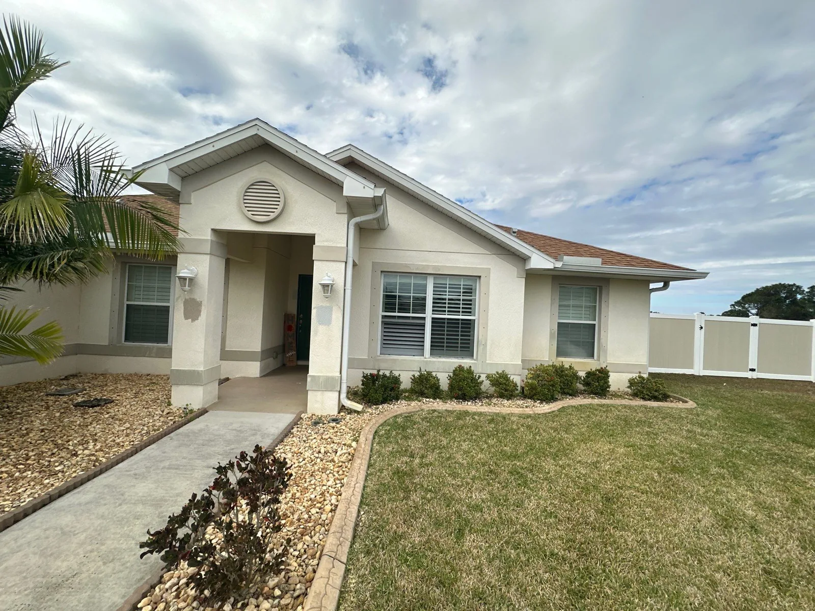 Exterior Painting in Port Saint Lucie, Florida. Front elevation of a single-storey house painted beige with white trim and gable entry, lawn and rock beds.