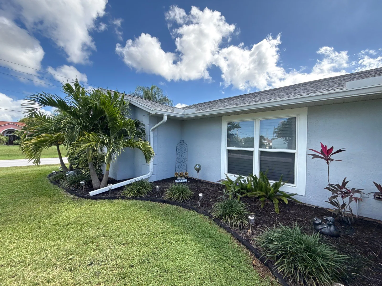 Exterior Painting in Port Saint Lucie, Florida. Corner of a single-storey light blue stucco house with white trim and fascia, planting bed with palms and a mown lawn.