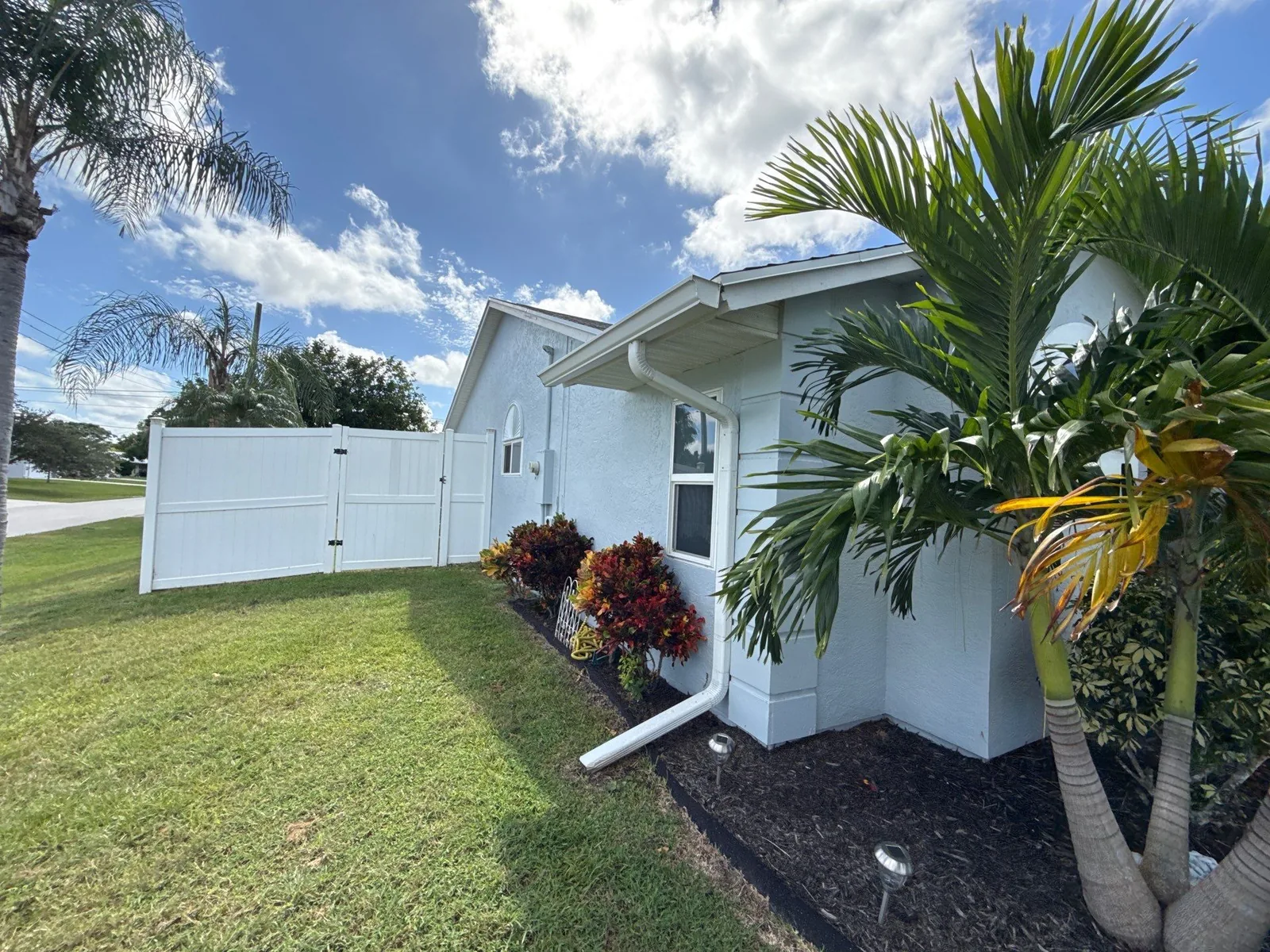 Exterior Painting in Port Saint Lucie, Florida. Side elevation of a white stucco single-storey house with white vinyl fence, downspout and lawn.