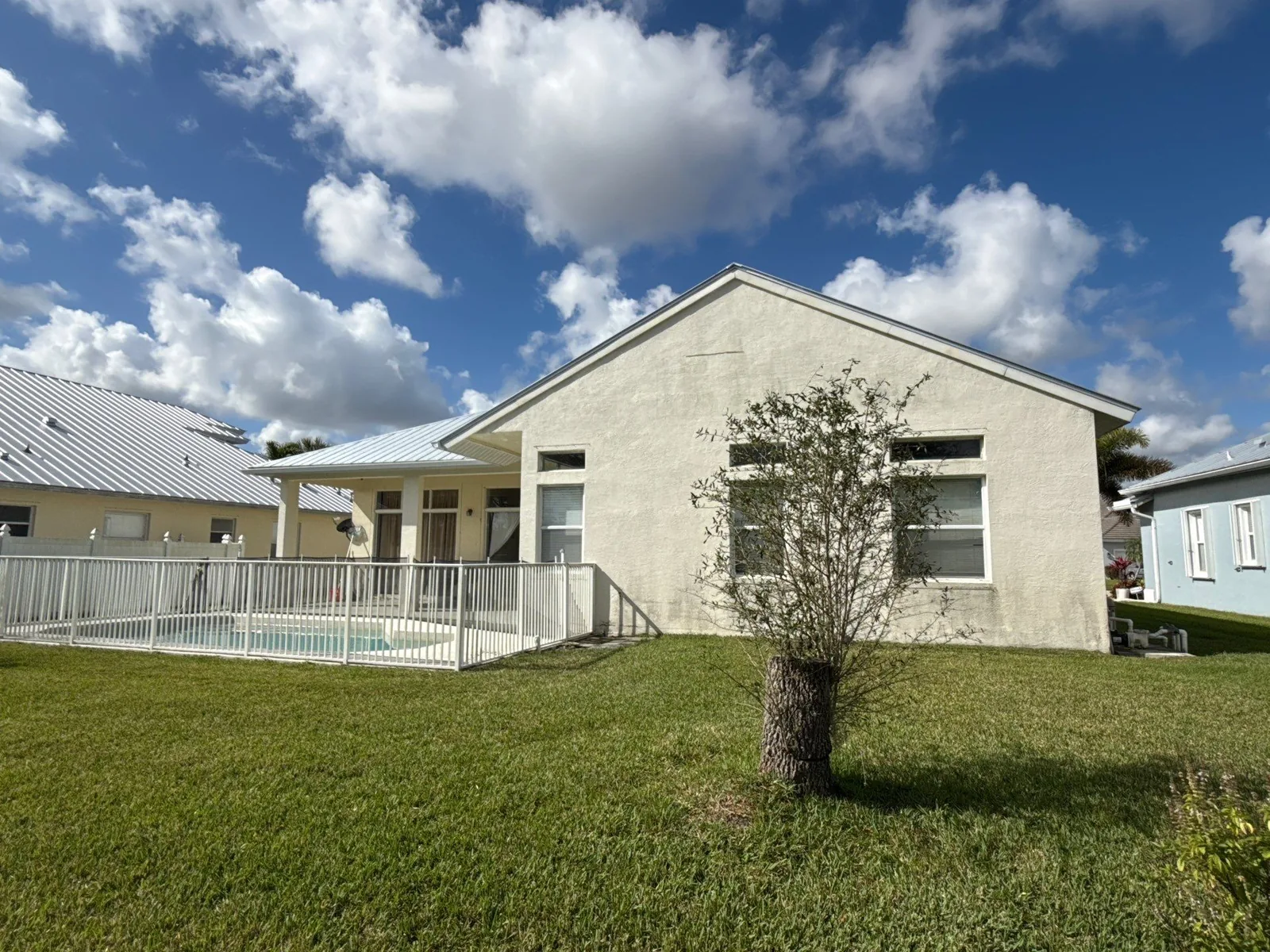 Exterior Painting in Port Saint Lucie, Florida. Rear corner of a cream stucco single-storey house with a metal roof, fenced pool to one side and lawn in front.
