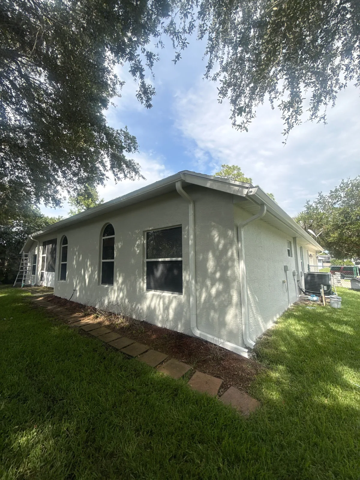 Exterior Painting in Port Saint Lucie, Florida. Single-storey stucco house painted white, shown from the corner with soffit and fascia, dark windows, a paver border and lawn.