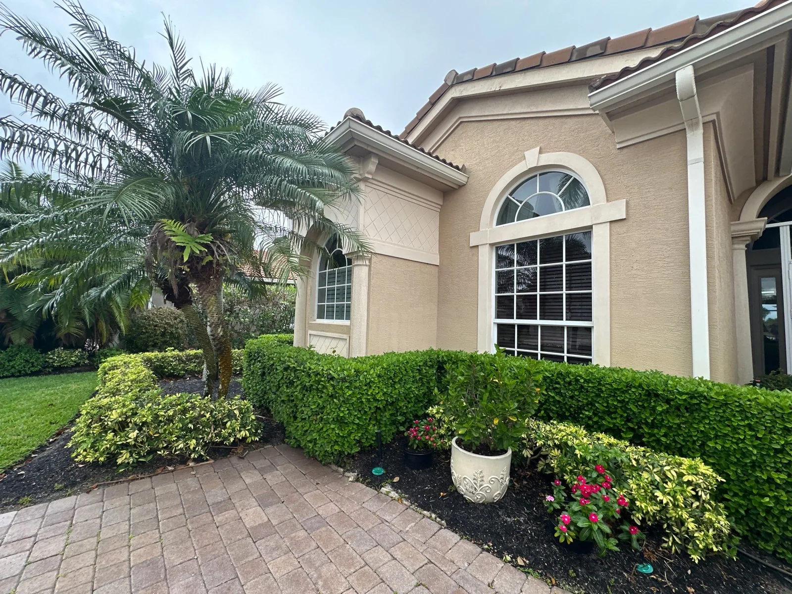 Exterior Painting in Port Saint Lucie, Florida. Front corner of a cream stucco home with white window trim, an arched window, tile roof, trimmed hedges and a paver walkway.