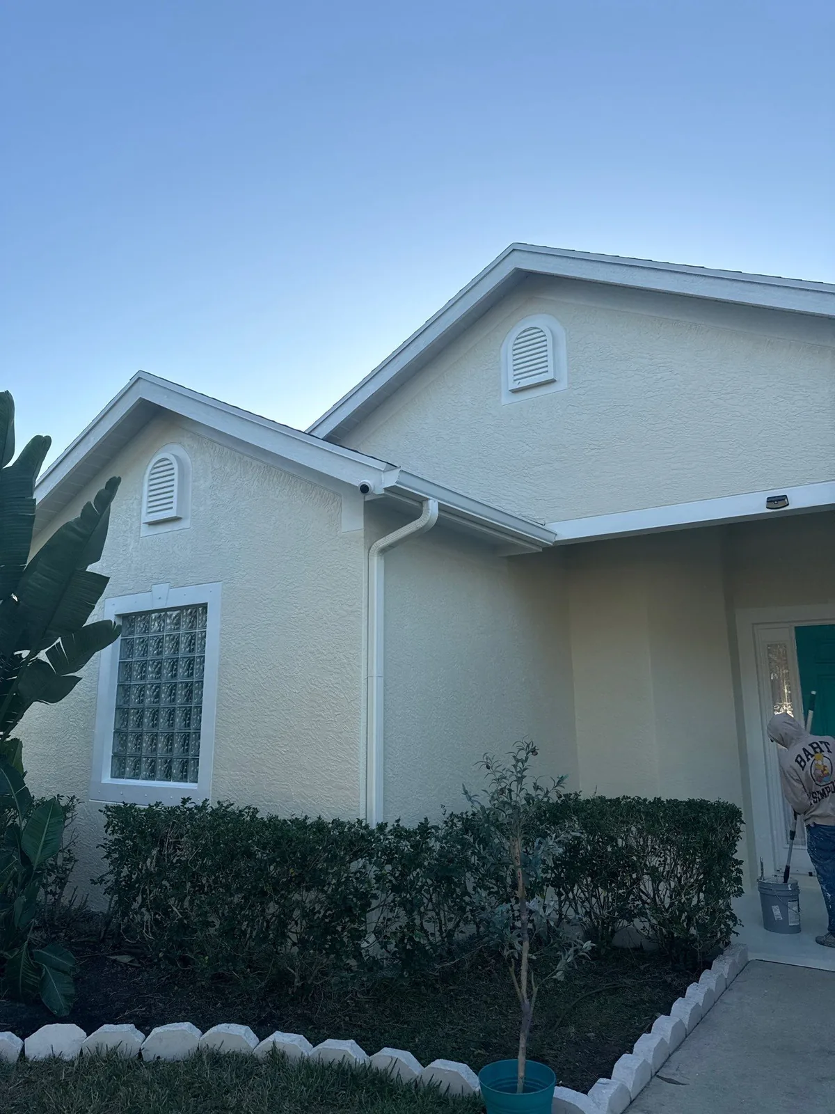 Exterior Painting in Port Saint Lucie, Florida. Pale yellow stucco house exterior with white fascia, gable vents and a glass block window above a trimmed hedge.