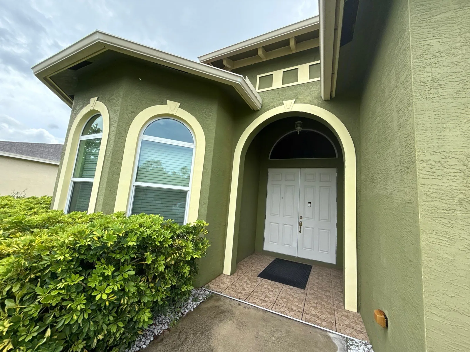 Exterior Painting in Port Saint Lucie, Florida. Front entry of a green stucco home with cream trim, arched entryway and arched windows, white front door and tiled porch.