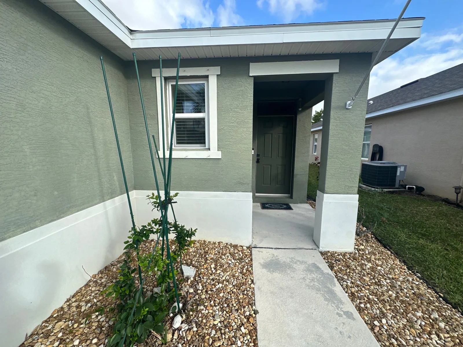 Exterior Painting in Port Saint Lucie, Florida. Covered front entry of a stucco home painted sage green with white window trim, white skirt band and a dark front door.