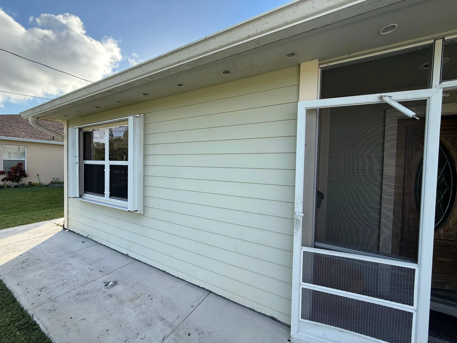 Exterior Painting in Port Saint Lucie, Florida. Pale green lap siding exterior wall with white window and screen door trim under a soffit.