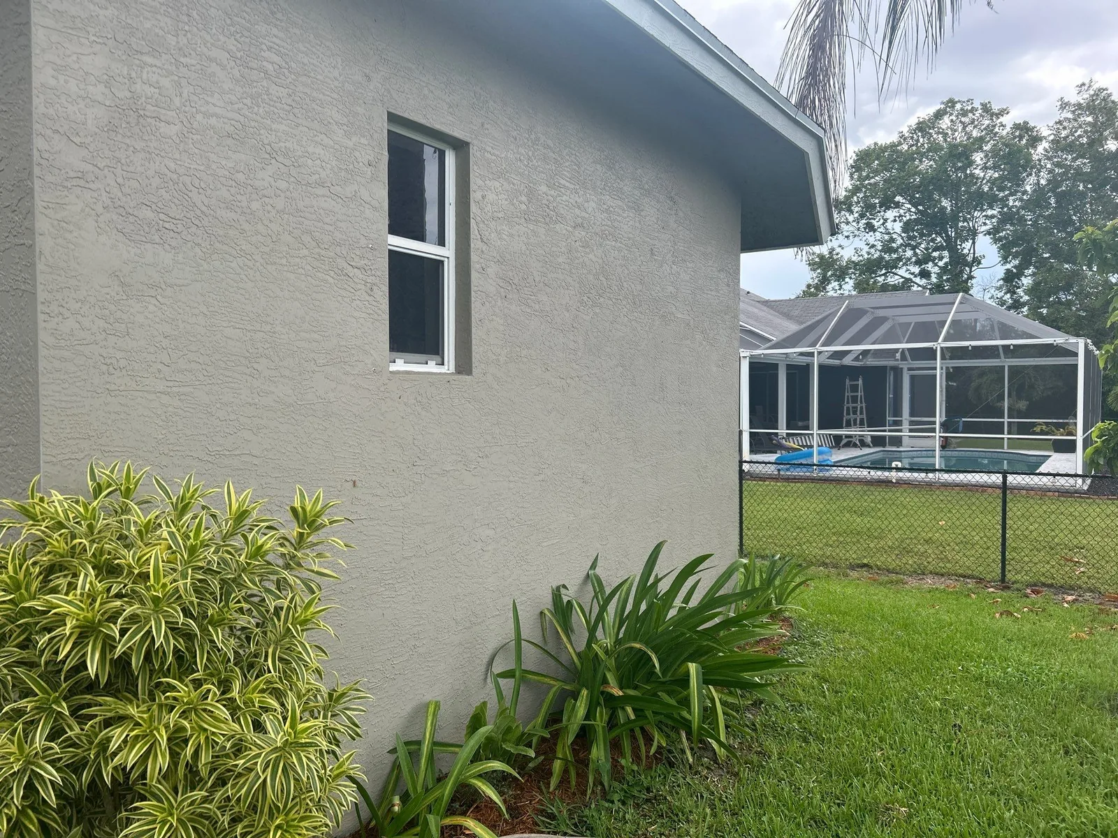 Exterior Painting in Port Saint Lucie, Florida. Corner of a single-storey house, grey stucco with a white-trimmed window, shrubs and lawn in front.