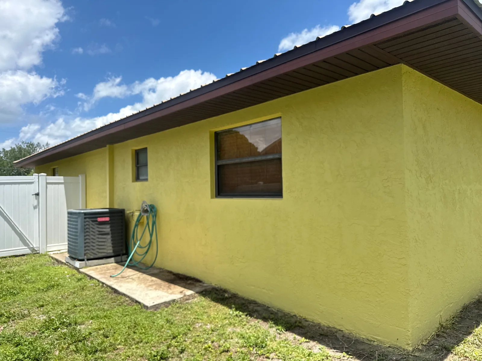 Exterior Painting in Port Saint Lucie, Florida. Side elevation of a single-storey stucco home painted yellow with dark window trim, AC condenser at the base of the wall.