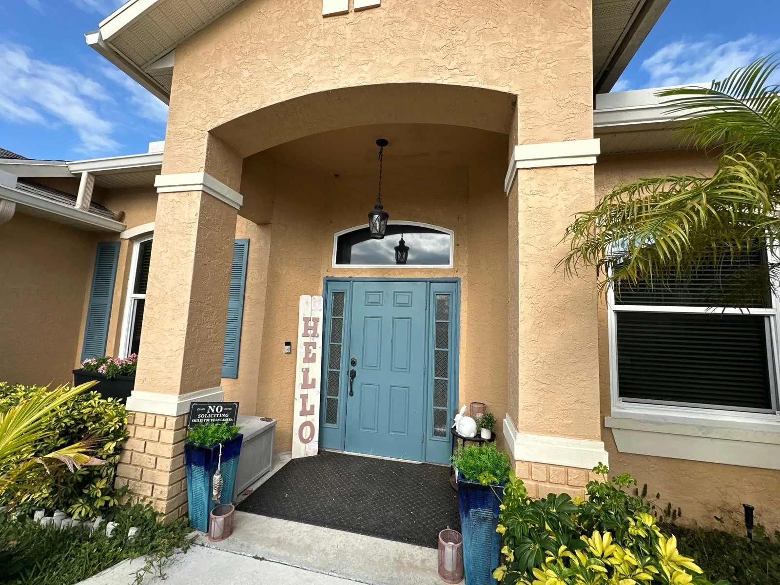Exterior Painting in Port Saint Lucie, Florida. Arched front entry of a tan stucco home with white trim, a blue-painted front door and matching shutters, and planters either side.